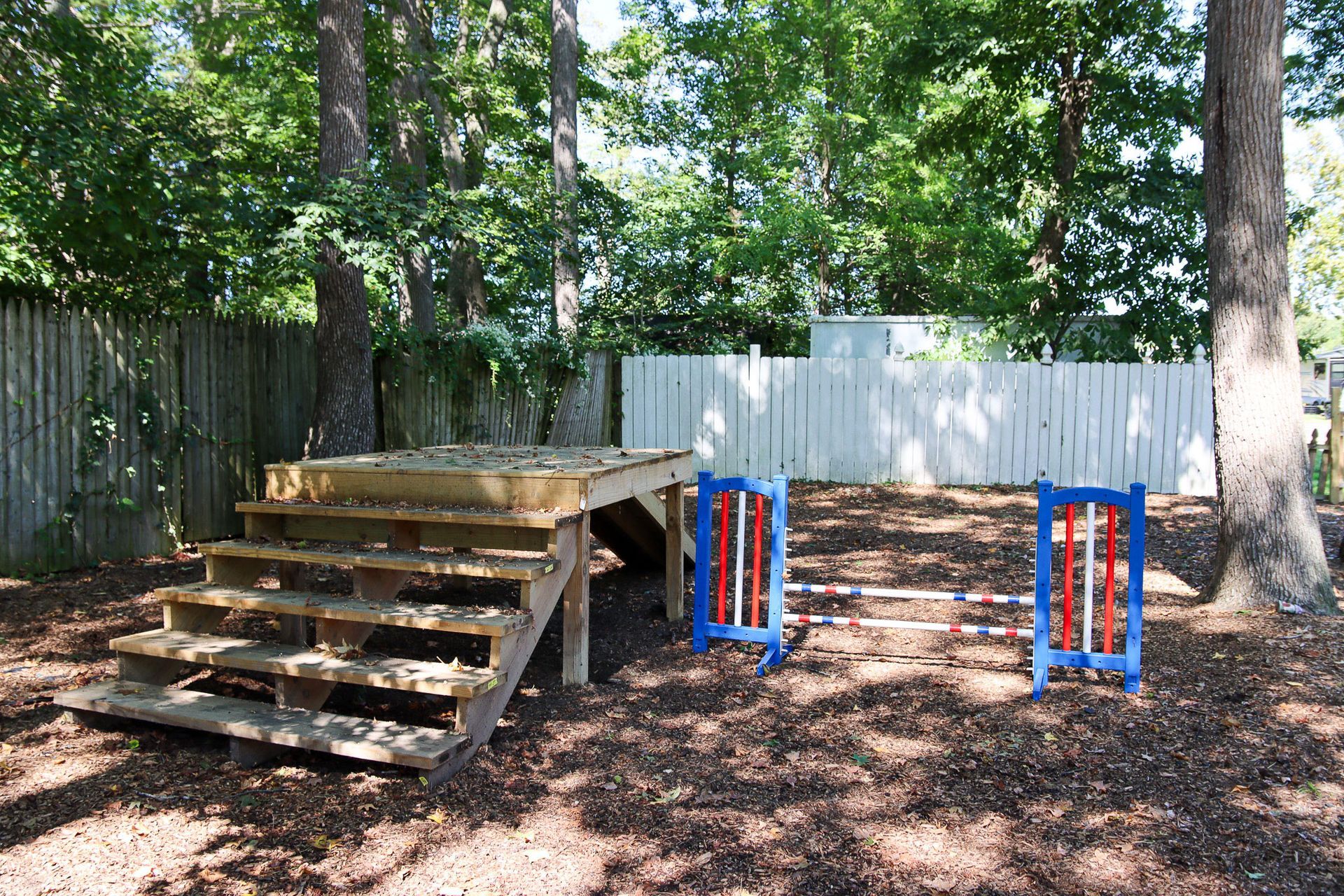 Backyard with a wooden platform, blue and orange stepping obstacles, and trees behind a white fence
