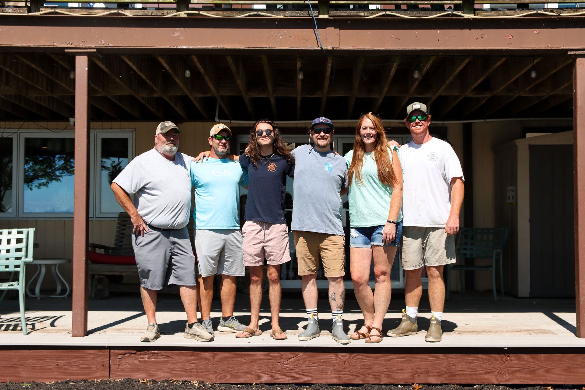 Six people standing side by side in front of a building, smiling in sunlight.