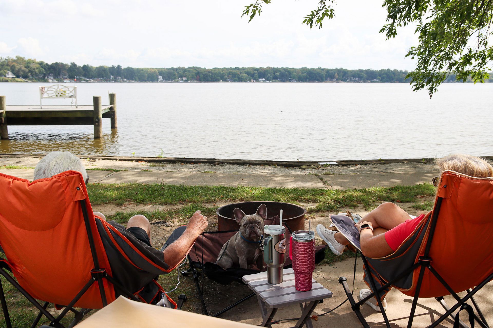 Two people relaxing in orange chairs by a lakeside fire pit, with a dock and calm water in the background