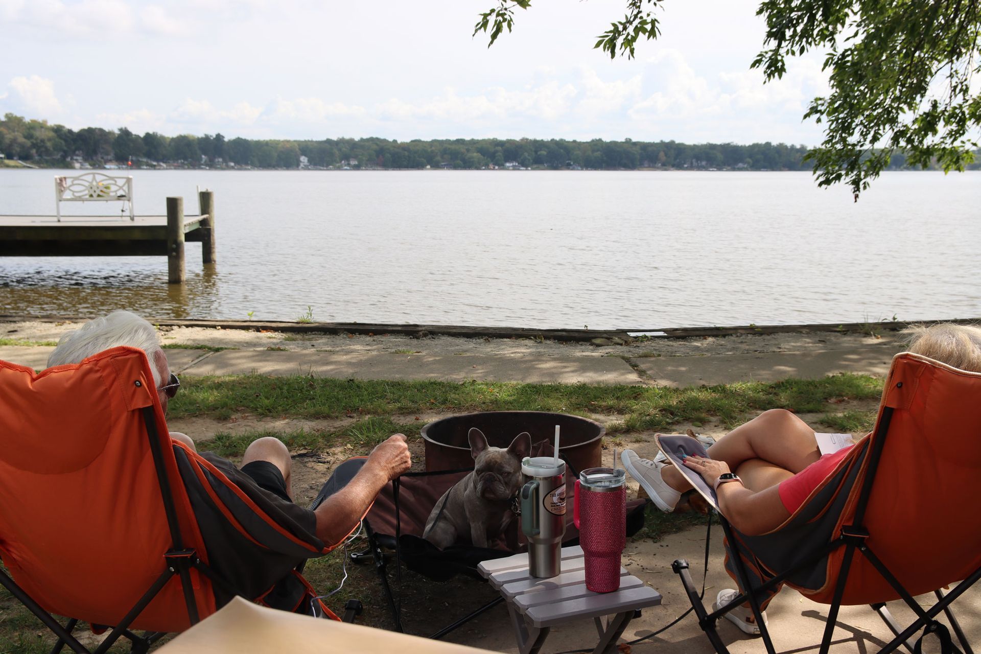 Two people relaxing in orange chairs by a lakeside, with drinks on a table and a calm water view.