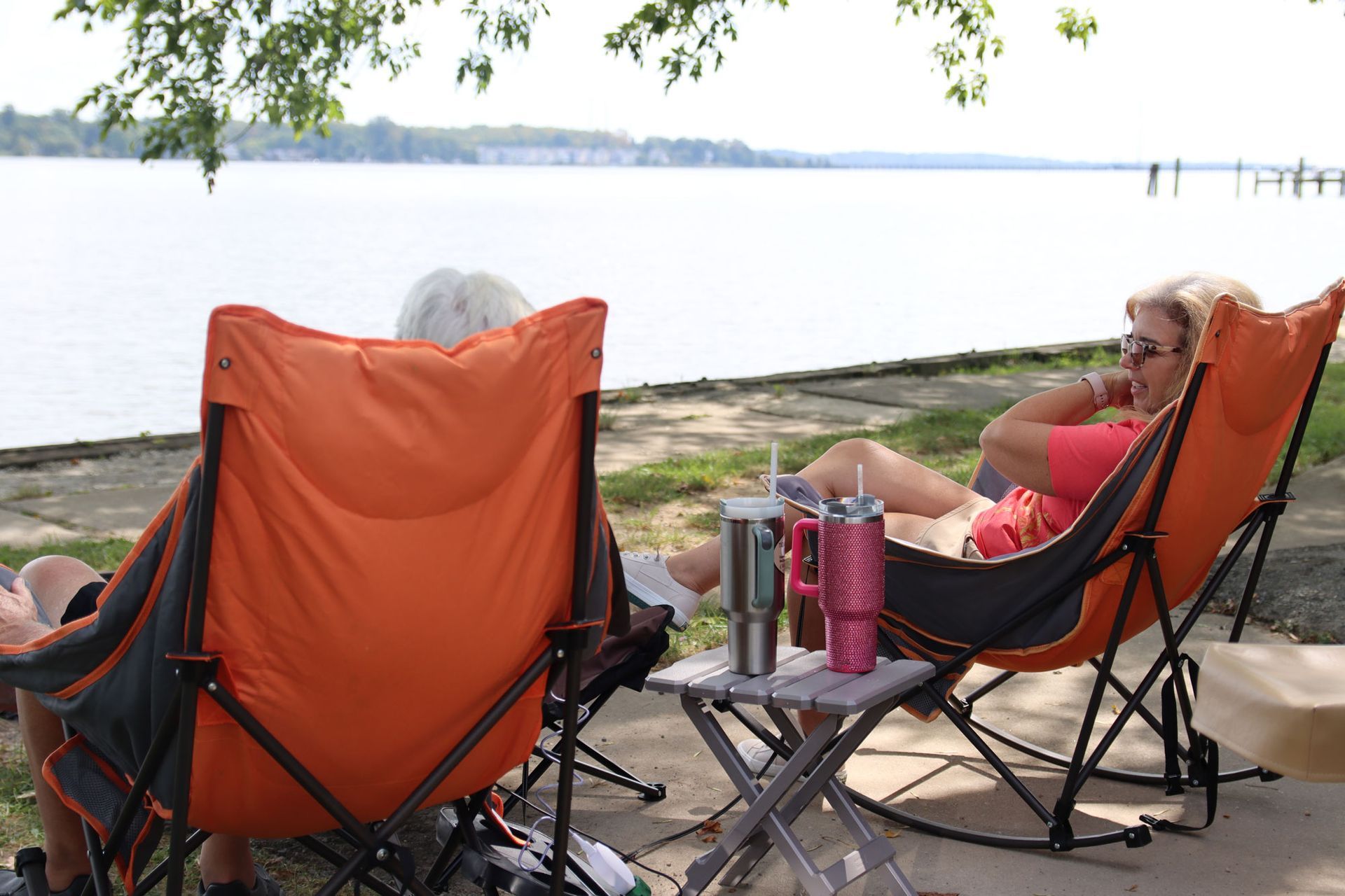 People relaxing in orange camping chairs beside a calm lake, with drinks and trees overhead