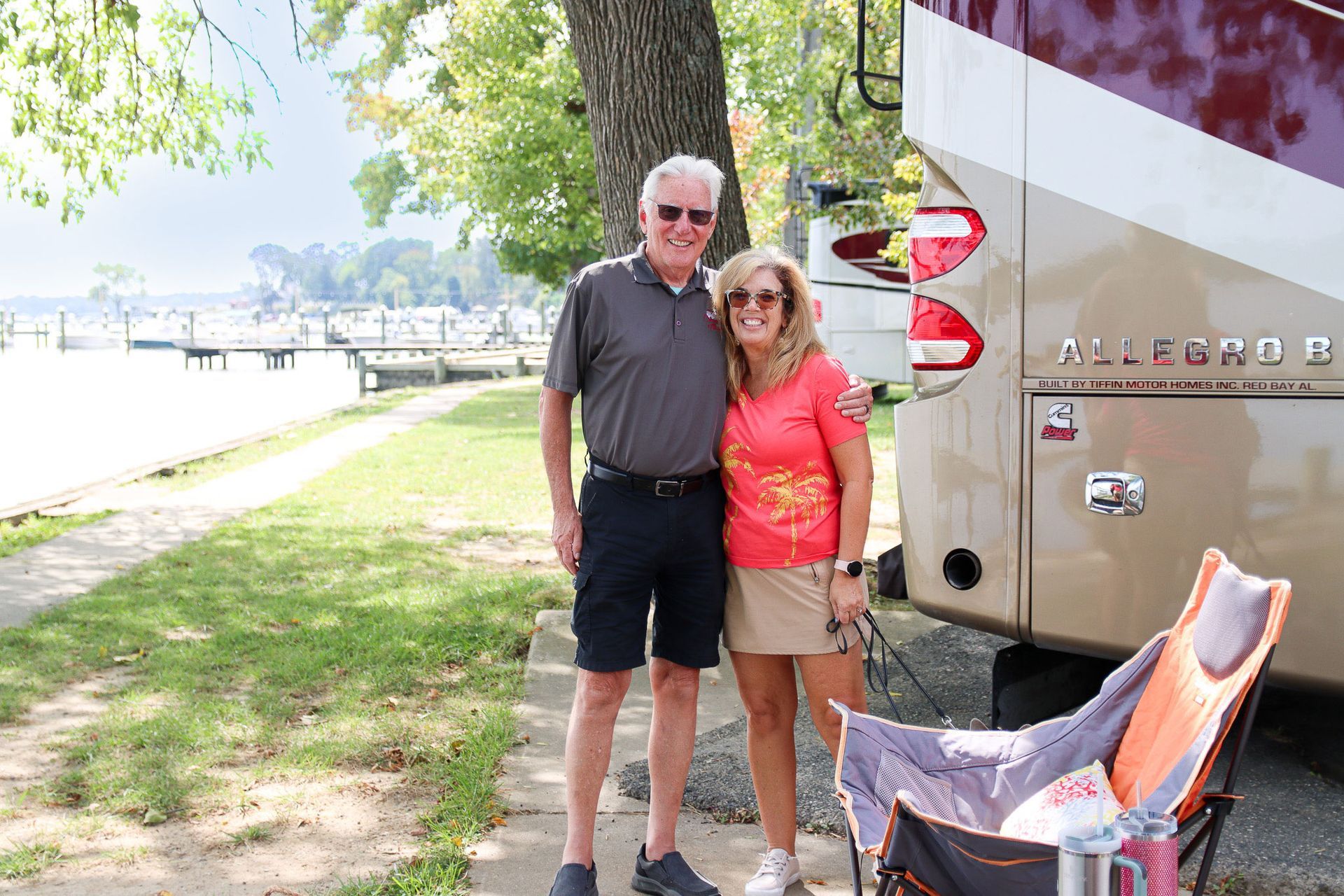 Two people standing by a lakeside RV, with a tree, lawn, and dock in the background.
