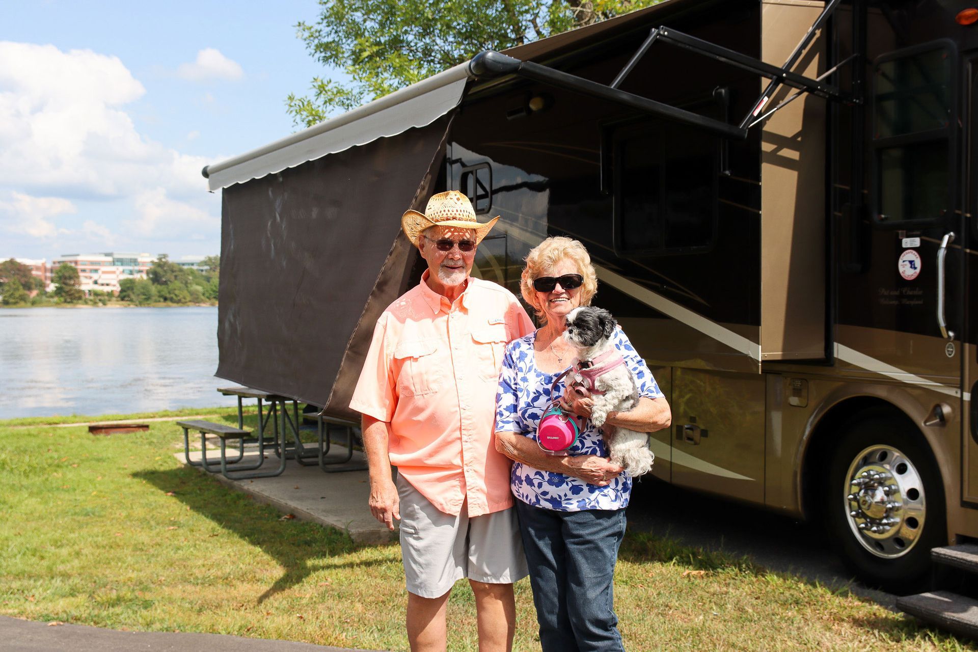 Two people standing beside a black RV by a lake on a sunny day