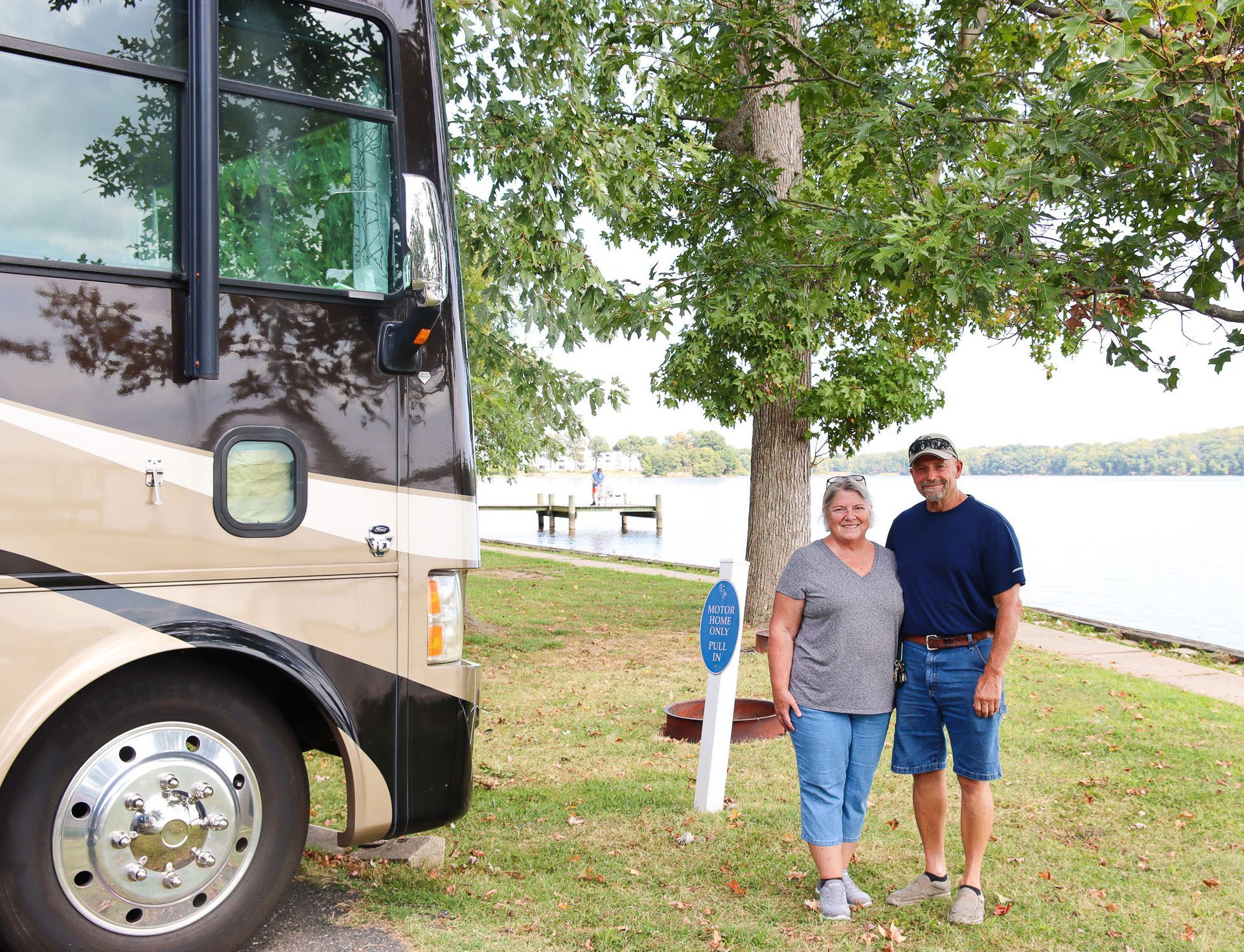 Two people standing beside a parked RV near a lakeside tree and picnic area