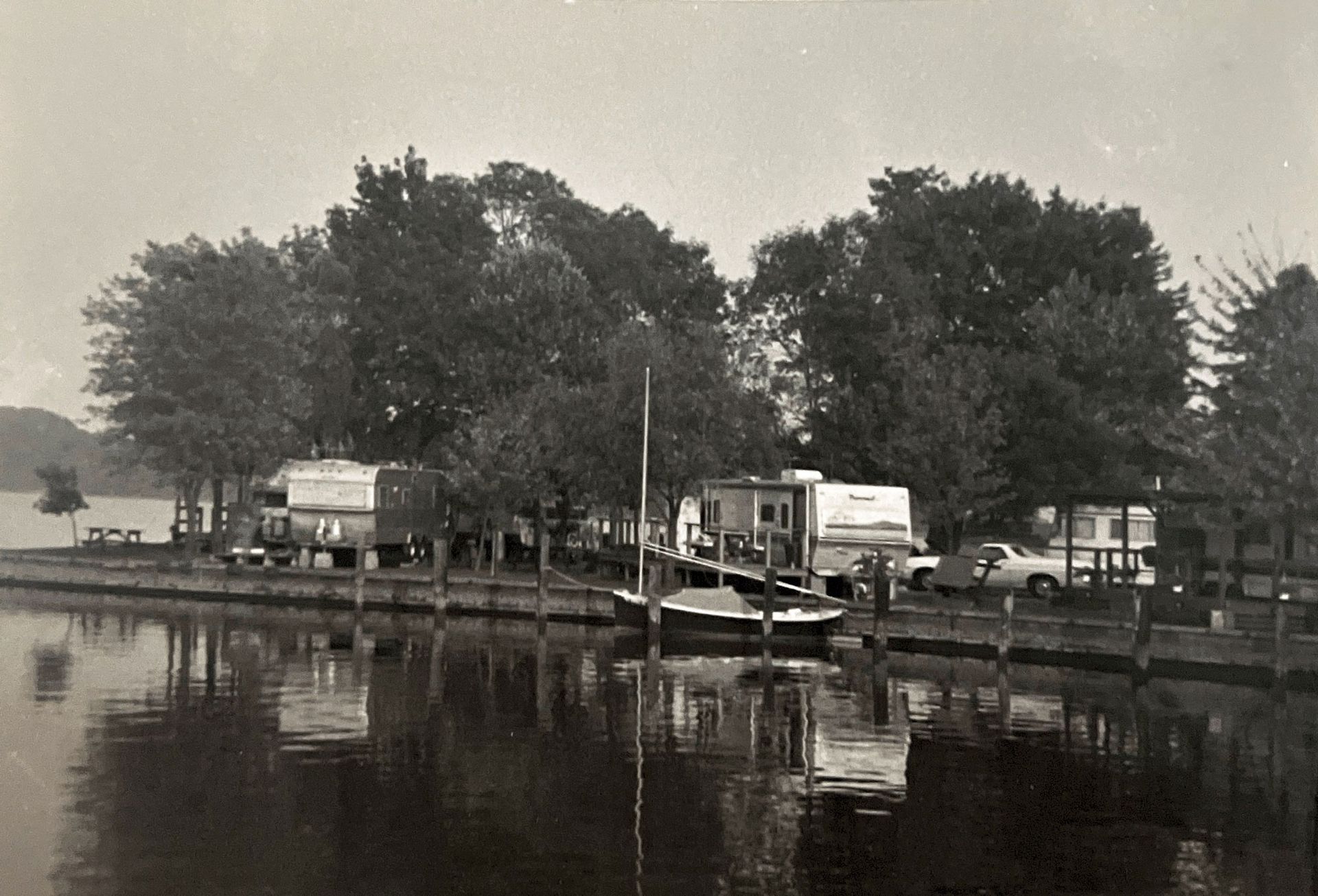 Black-and-white lakeside marina with boats, trees, and parked trucks reflected in calm water