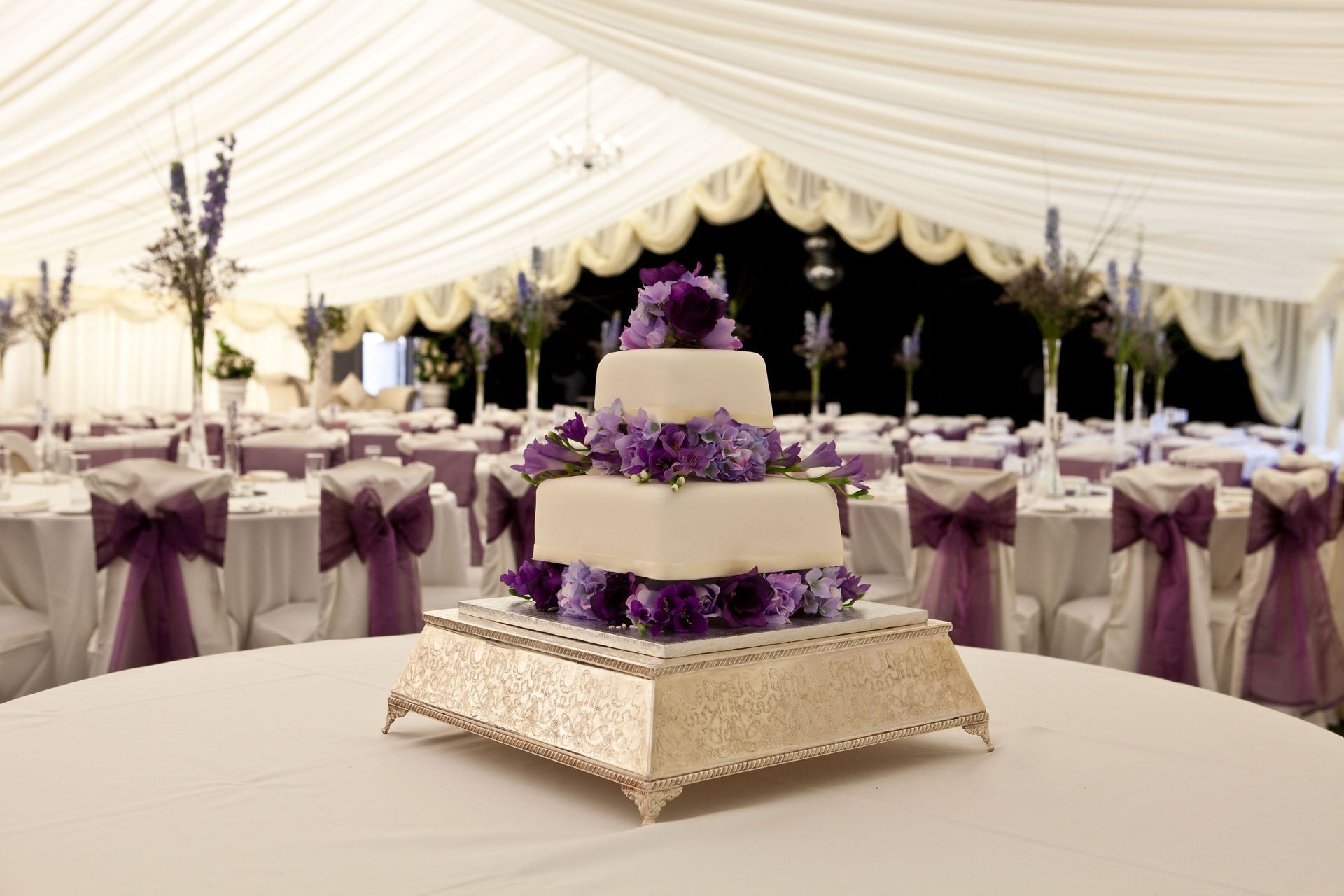 Purple-and-white tiered cake on a decorated table inside a white event tent, with matching table settings behind it