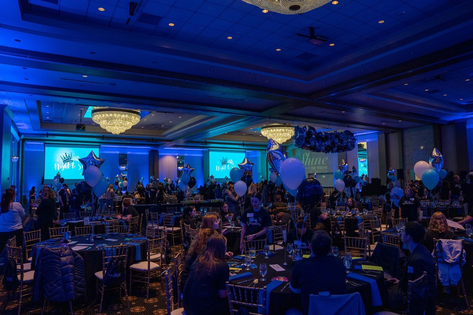 Blue-lit banquet hall with round tables, guests seated, and stage lights at an event