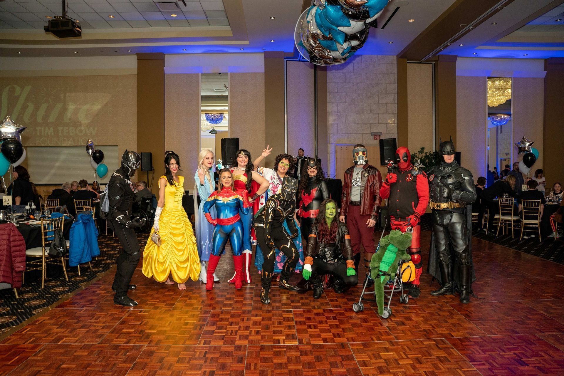 Costumed superhero group posing in a banquet hall with balloons and a dance floor