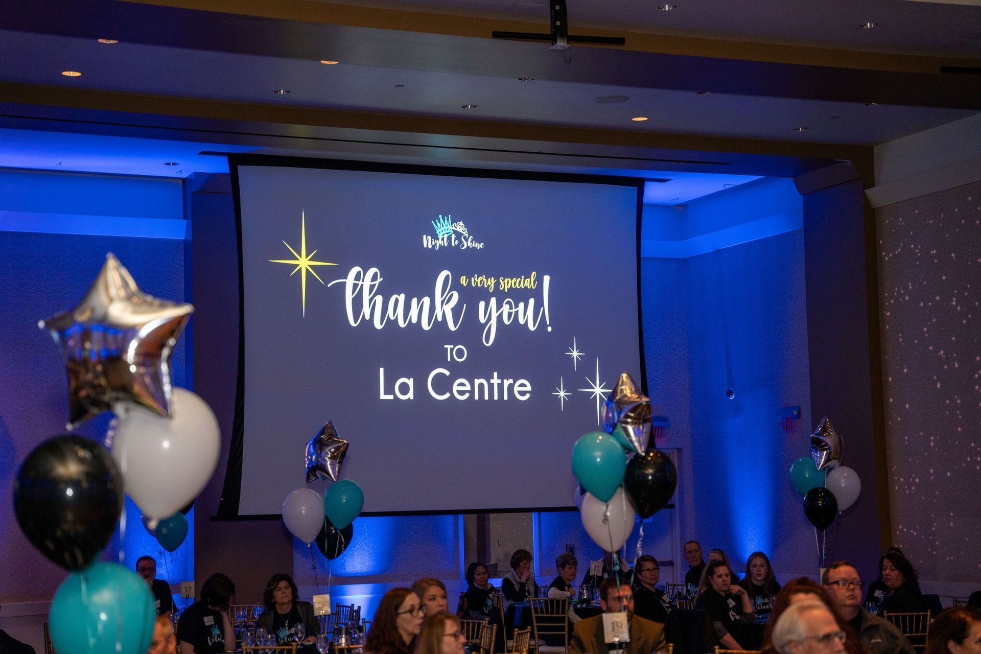 Blue-lit event stage with “Thank you! La Centre” on screen and balloon decorations over a seated audience