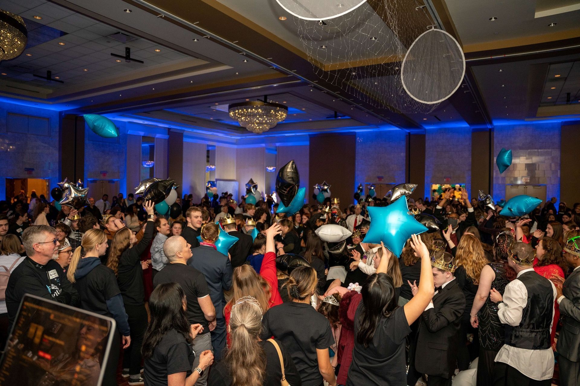 Crowded ballroom dance party with blue lighting, people in costumes, and star-shaped balloons