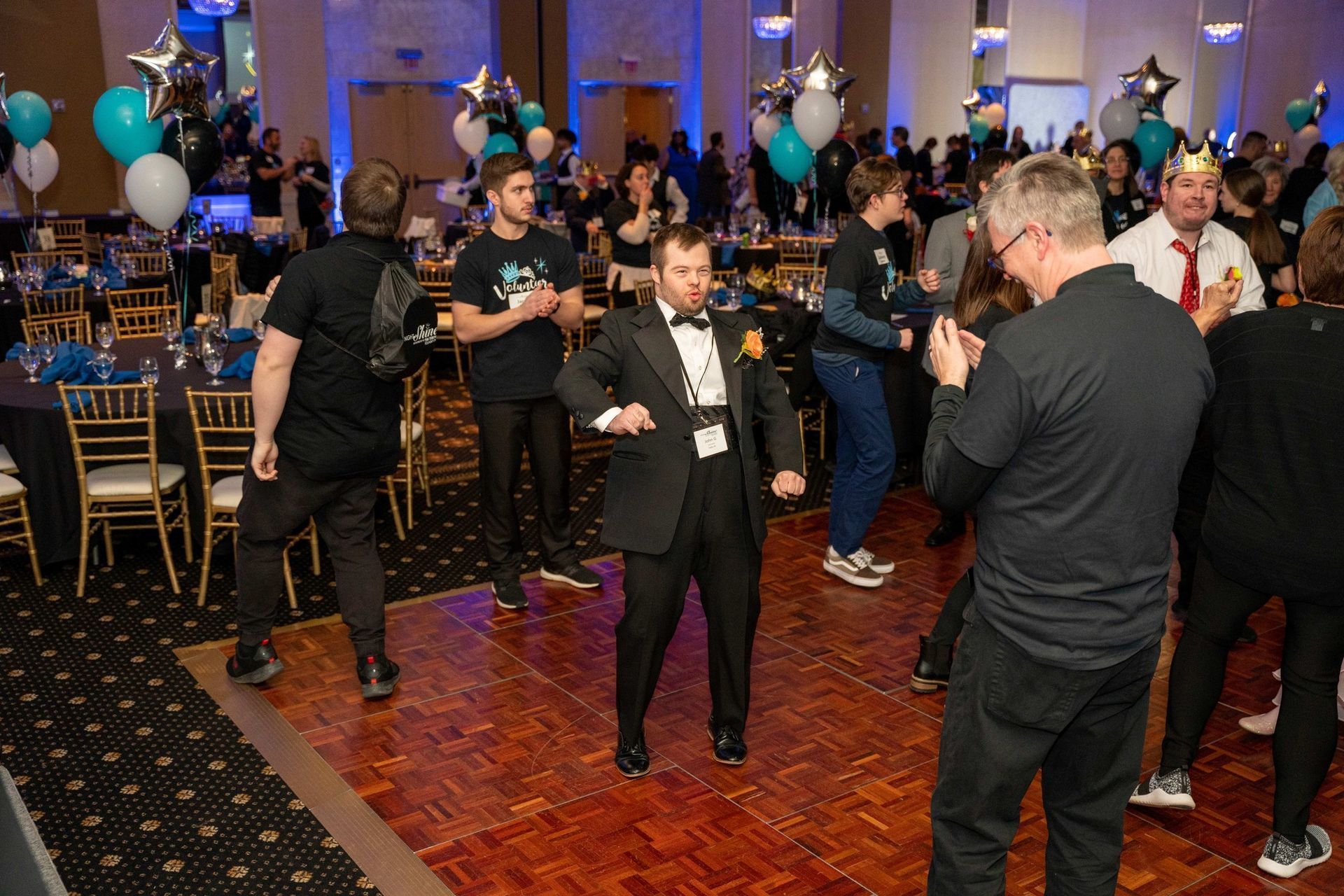 People mingling at a formal indoor event, with blue-lit balloons and gold chairs in the background.