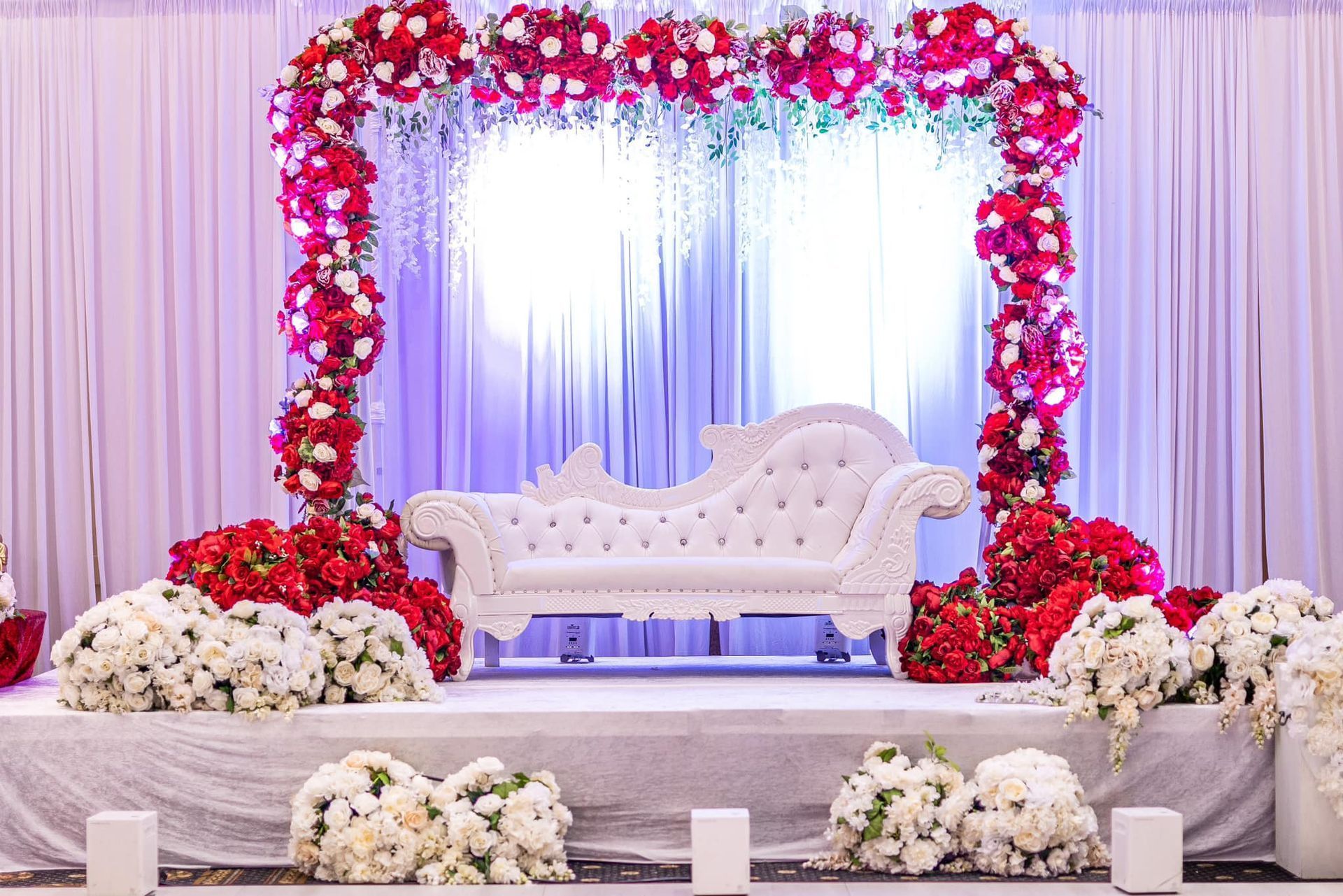 White sofa on a flower-decorated wedding stage with pink, red, and white floral arch backdrop