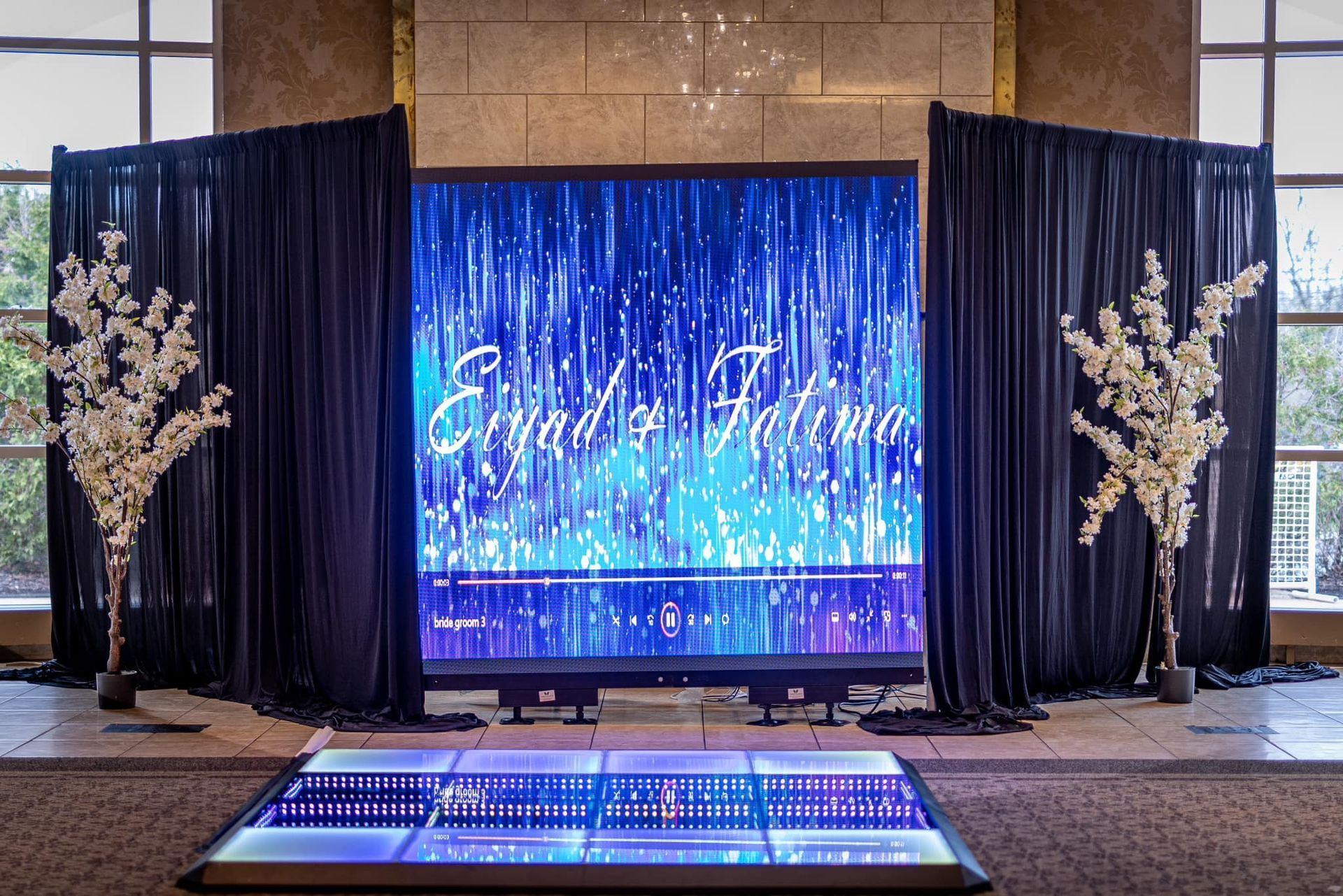 Elegant event stage with blue LED screen, black drapes, and floral arrangements at the sides