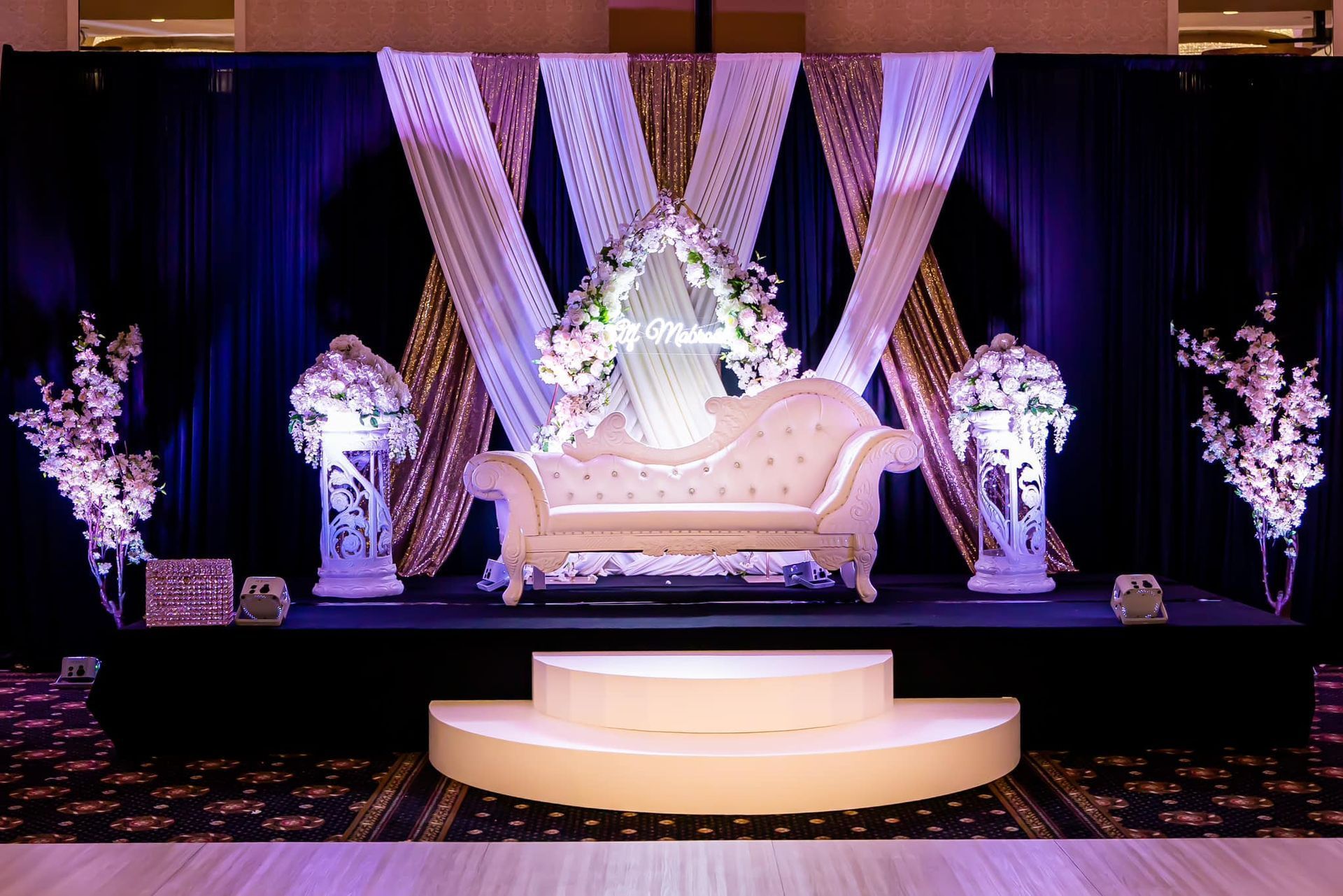 Lavishly decorated wedding stage with white sofa, draped lights, floral arrangements, and a lit circular platform