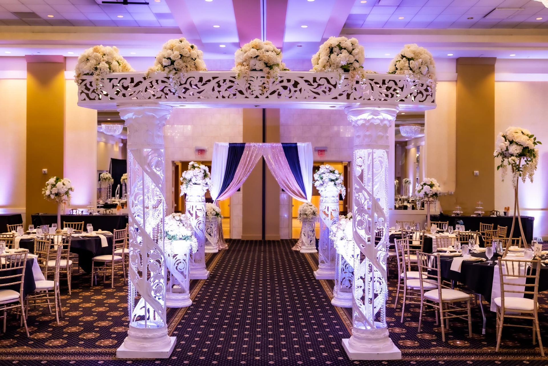 Decorated wedding reception aisle with a floral arch, purple drapes, and banquet tables in a ballroom