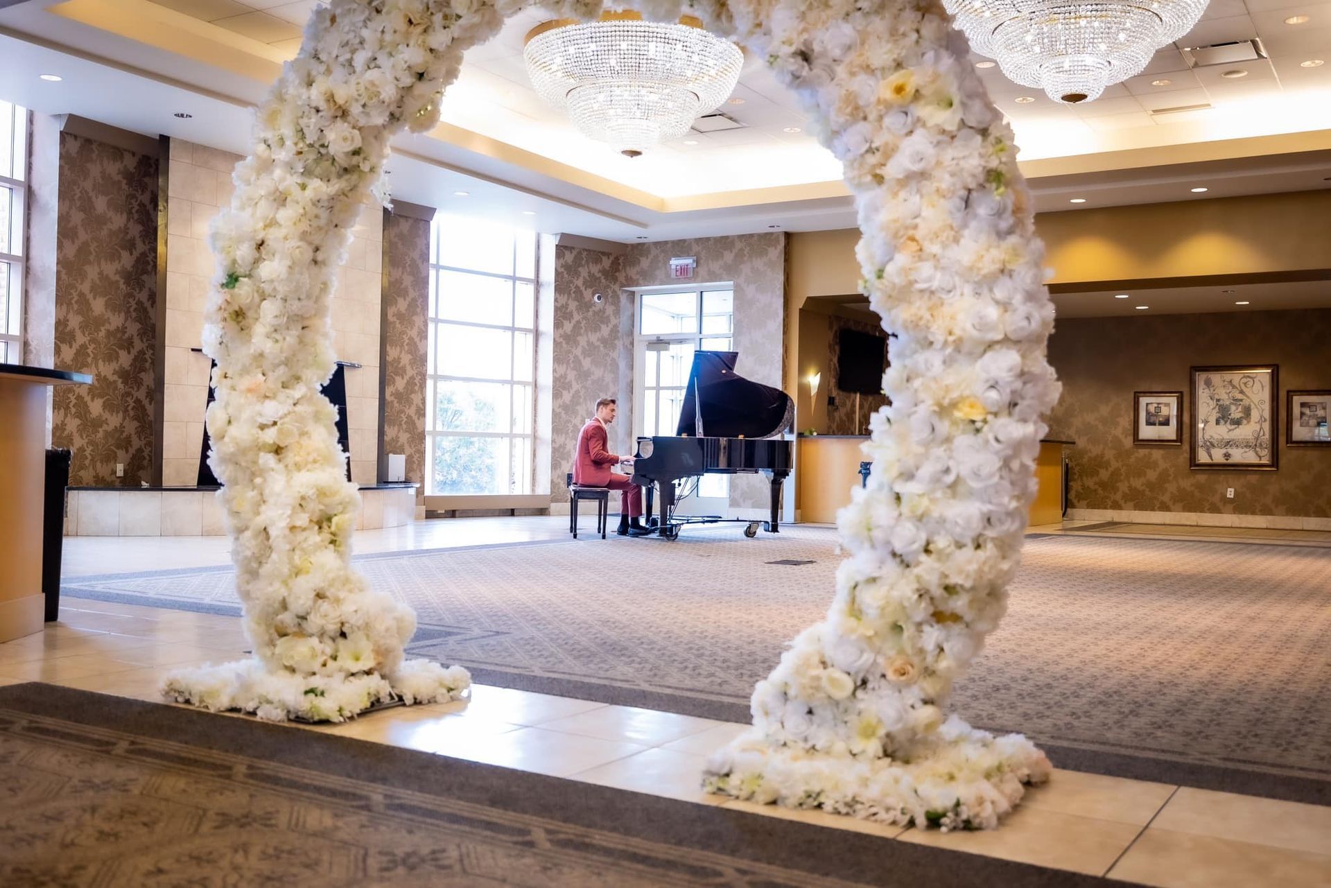 Flower arch in a hotel lobby with a grand piano and chandelier-lit ballroom decor