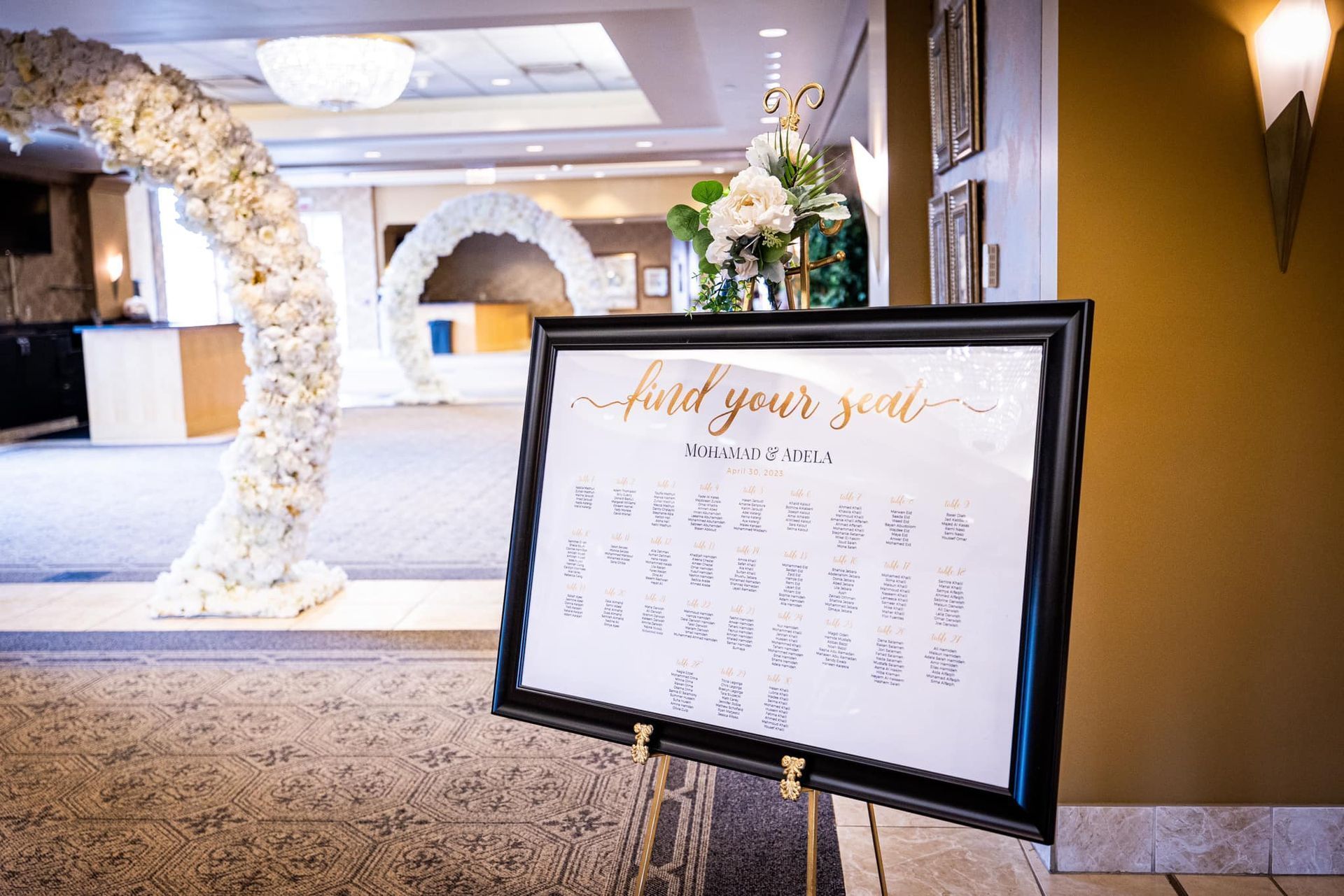 Wedding seating chart on a black easel beside a floral arch in a decorated venue hall