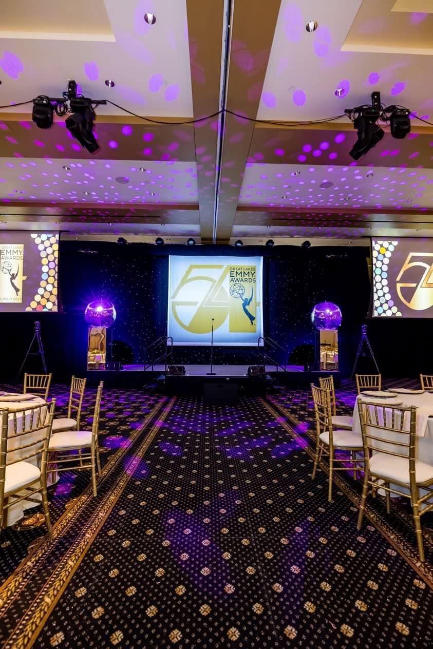 Elegant event hall with purple lighting, a stage, and banquet chairs arranged along a carpeted aisle.