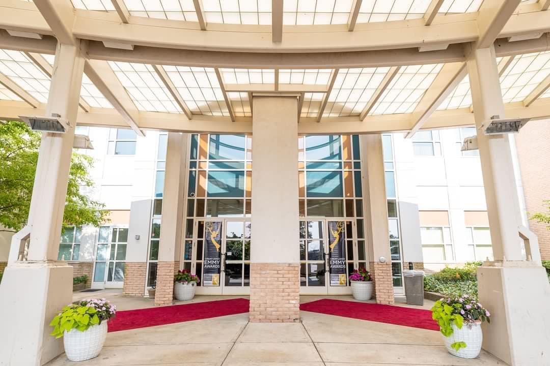 Covered hotel entrance with glass doors, white columns, and potted plants under a beige canopy