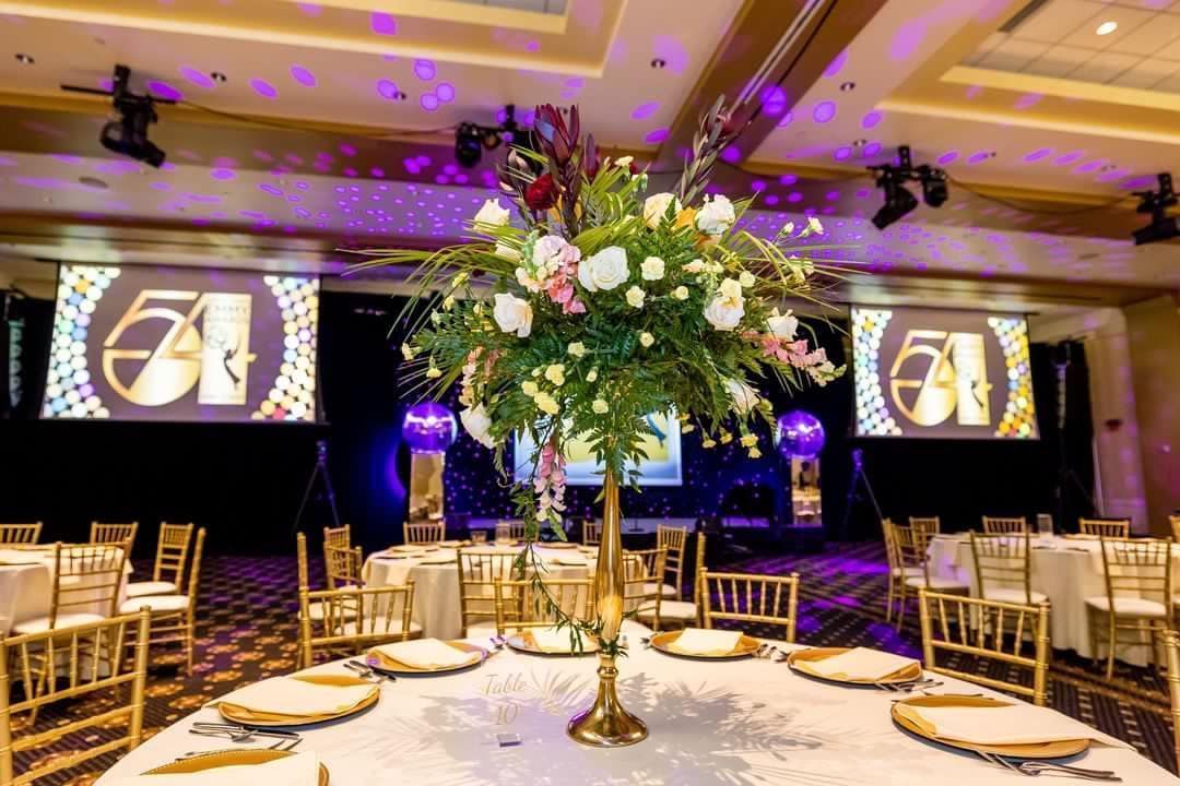 Elegant banquet hall with gold chairs, floral centerpieces, and projection screens displaying a logo under purple lighting