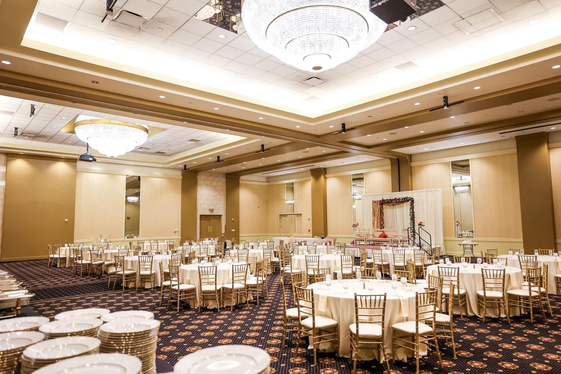 Elegant banquet hall with round tables, white chairs, and large chandeliers under warm lighting.