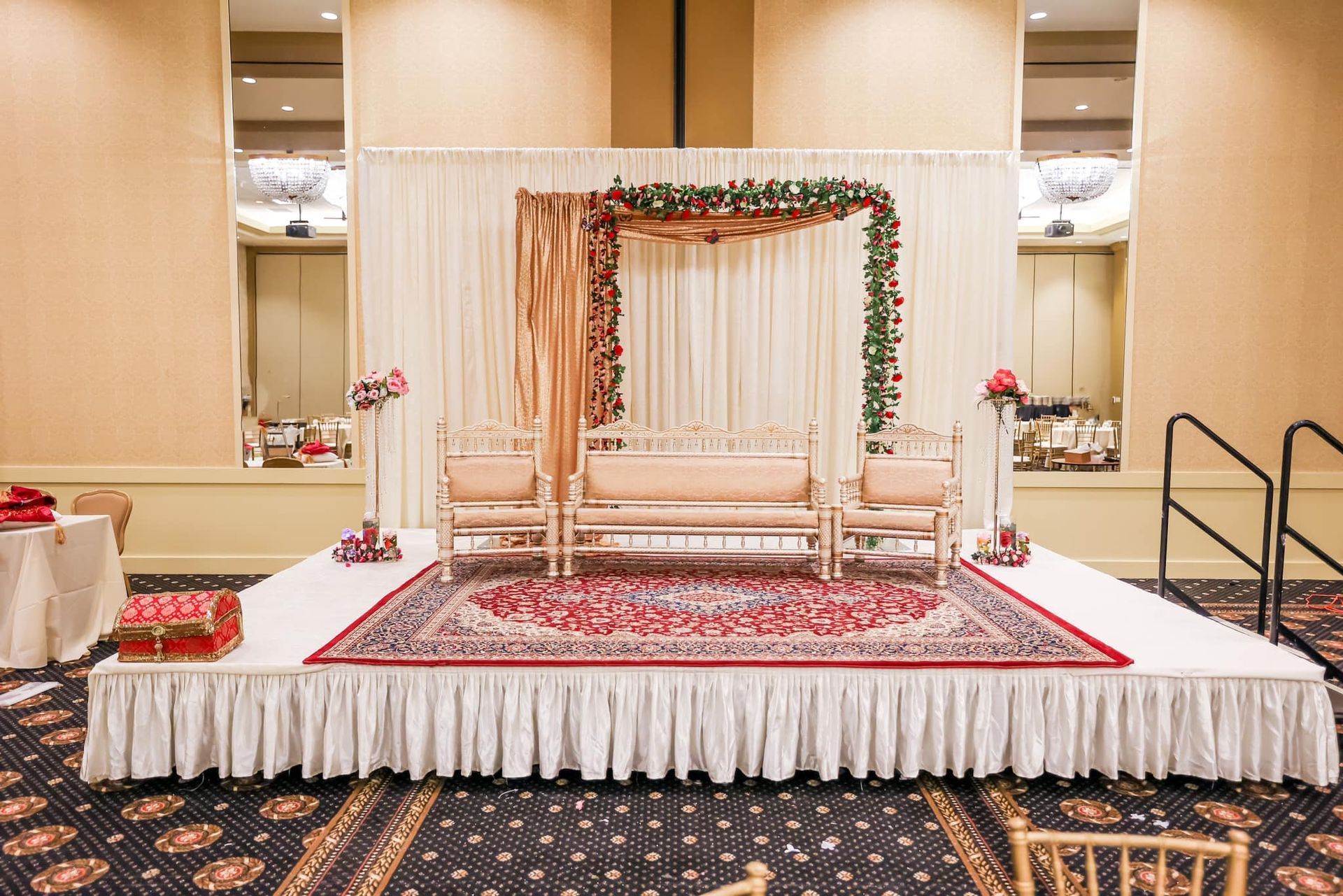 Decorated wedding stage with white sofas, floral arch, and red patterned carpet in a banquet hall