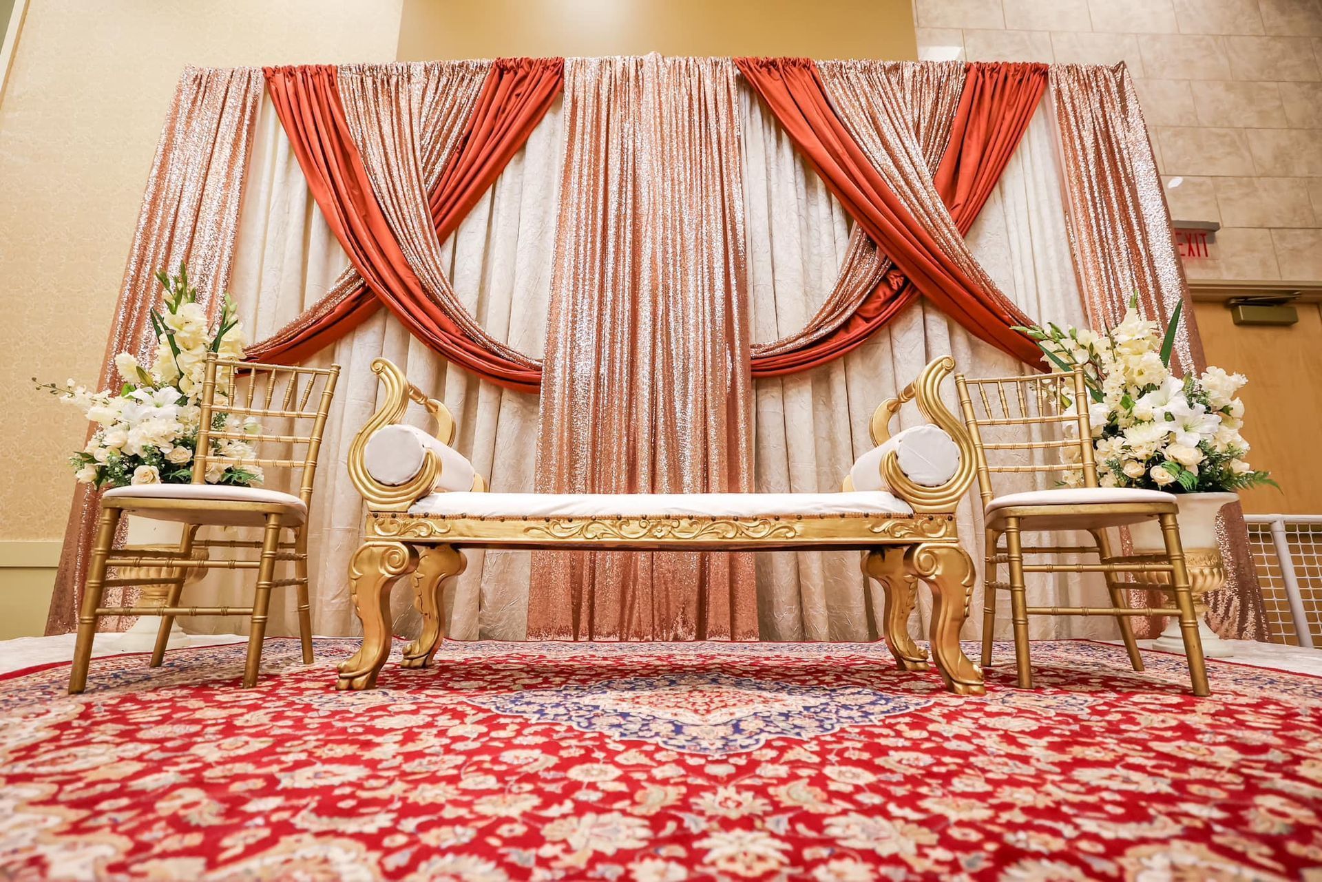 Decorated wedding stage with gold chairs, floral arrangements, draped curtains, and a red patterned carpet