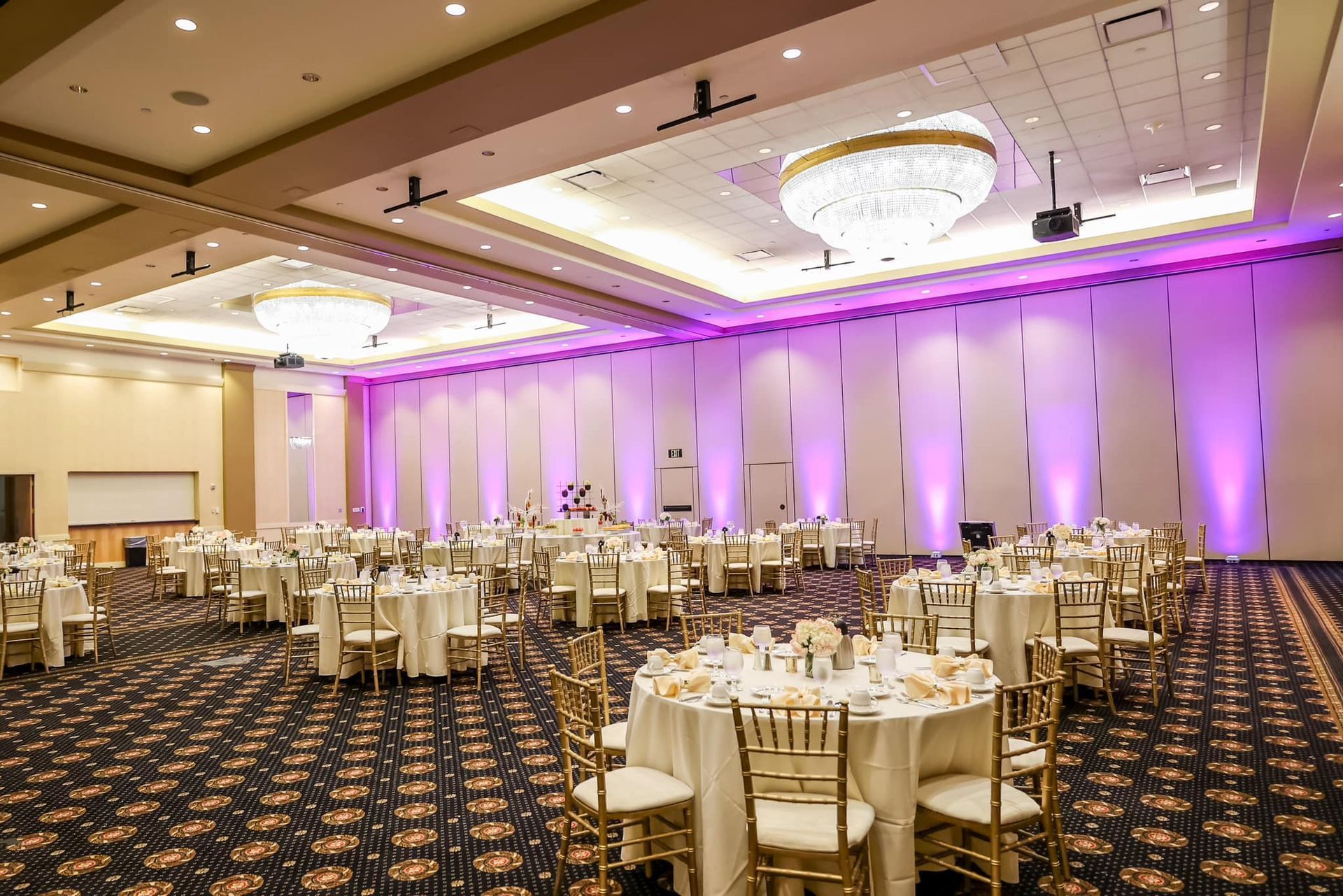 Elegant banquet hall with round tables, gold chairs, and purple uplighting under chandeliers.