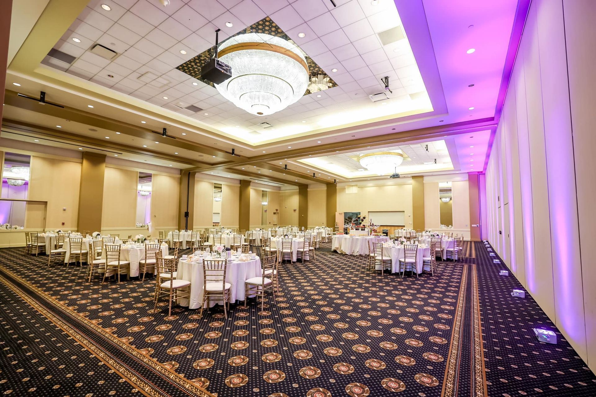 Elegant banquet hall with round tables, patterned carpet, chandeliers, and purple accent lighting
