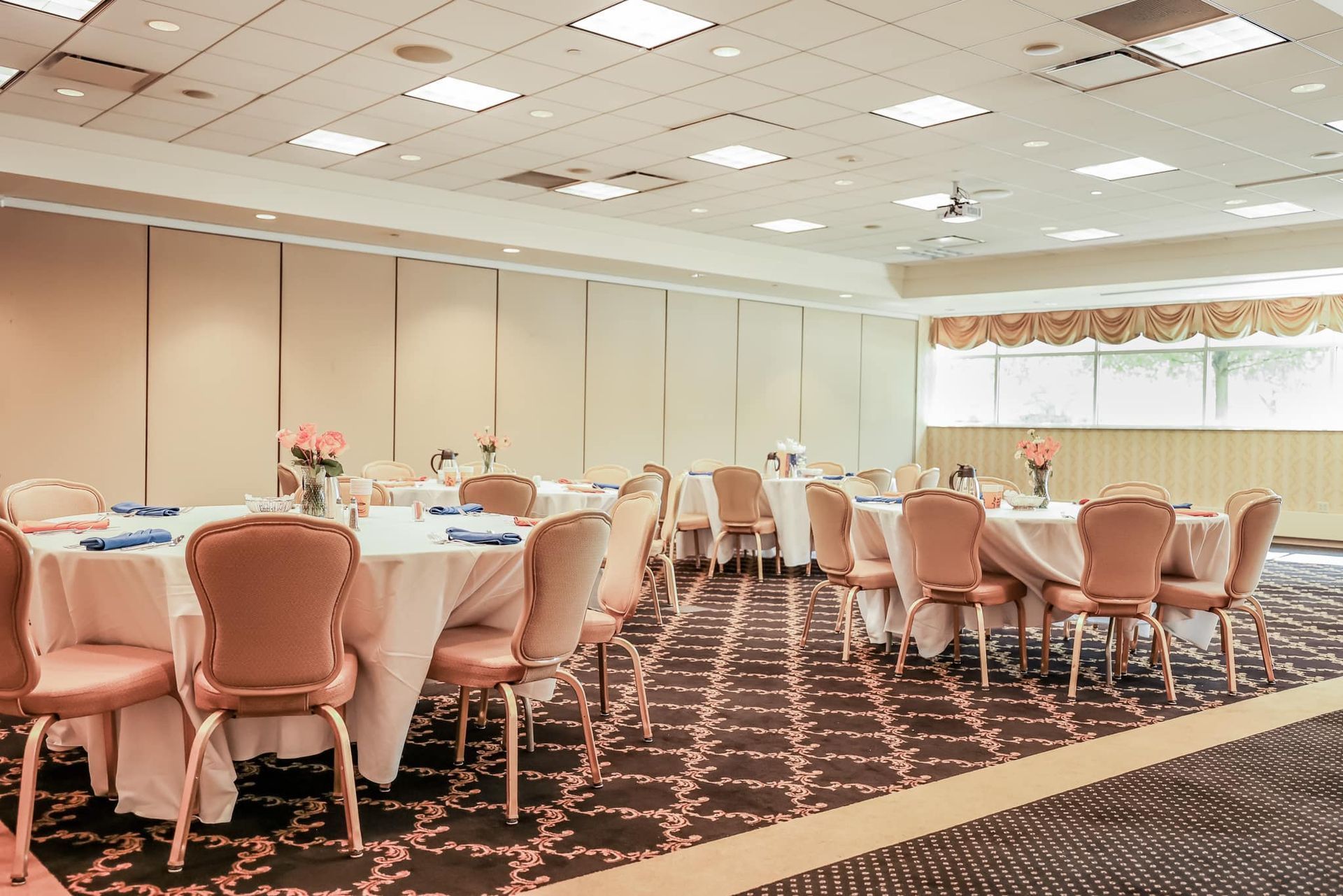 Empty banquet hall with round tables, pink chairs, and ceiling lights on patterned carpet