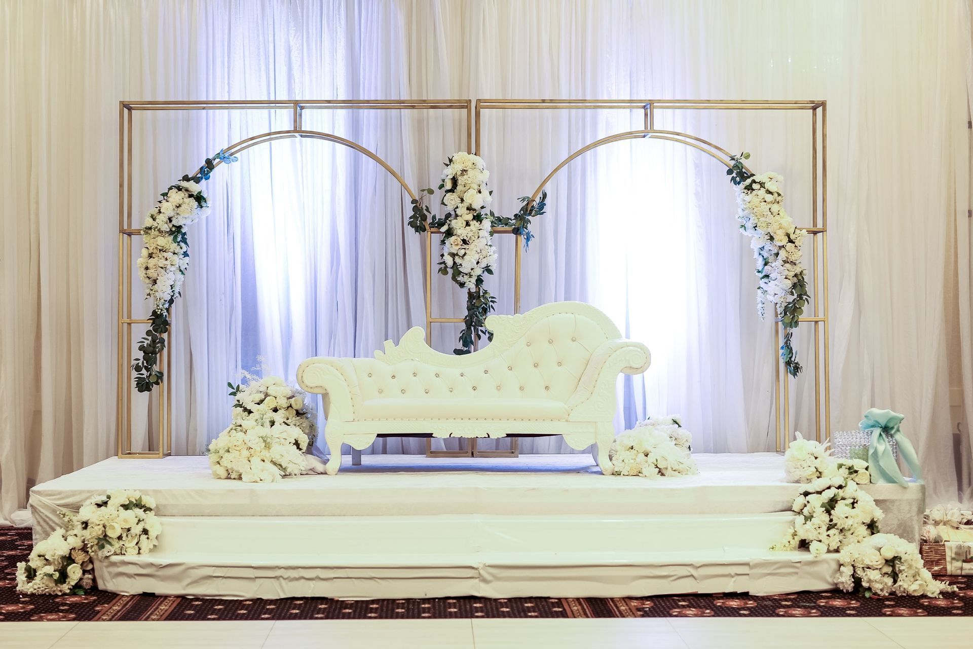 Elegant wedding stage with white floral arches, draped backdrop, and a white loveseat on a raised platform
