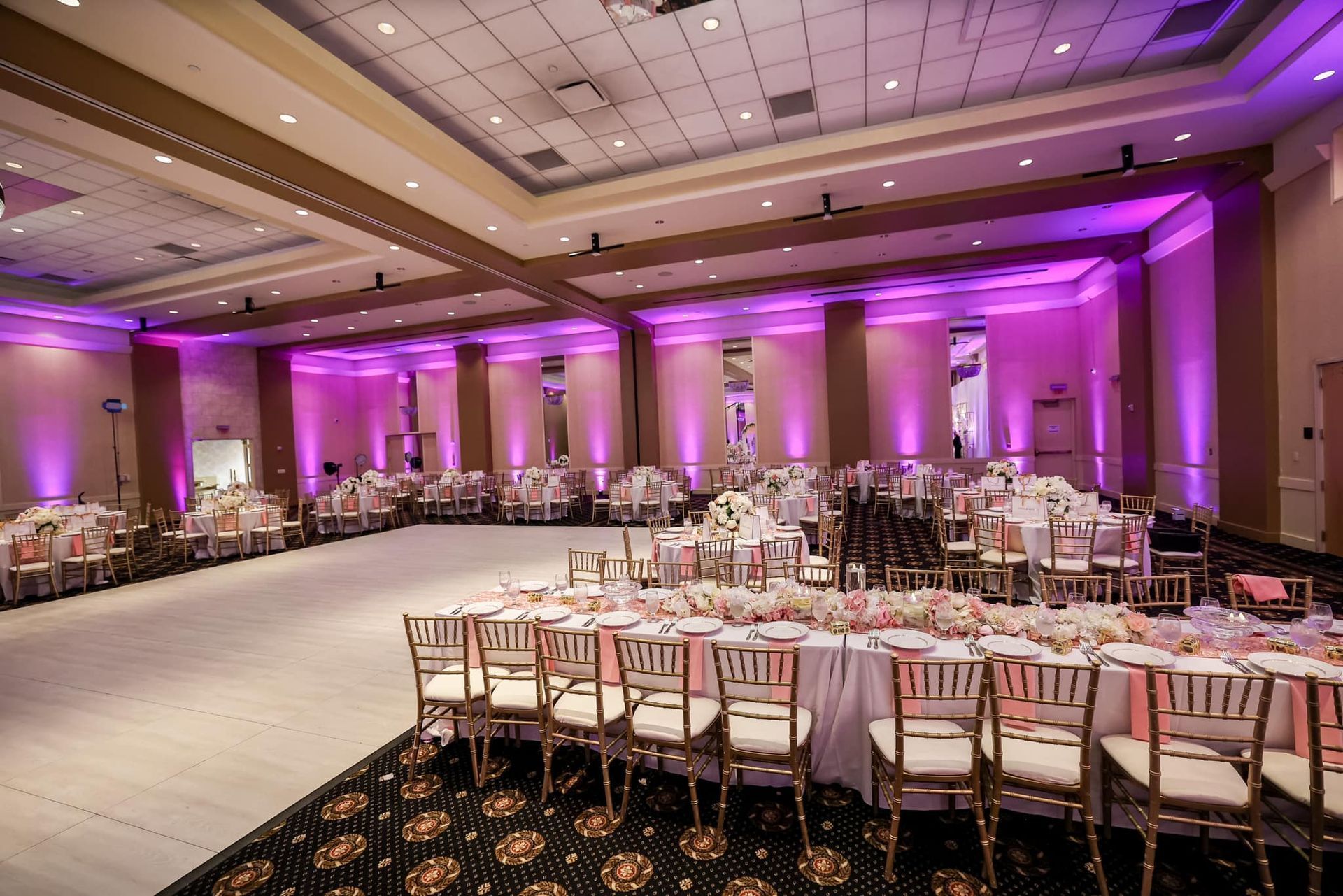 Empty banquet hall with round tables and white chairs, lit by purple uplighting and a carpeted dance floor