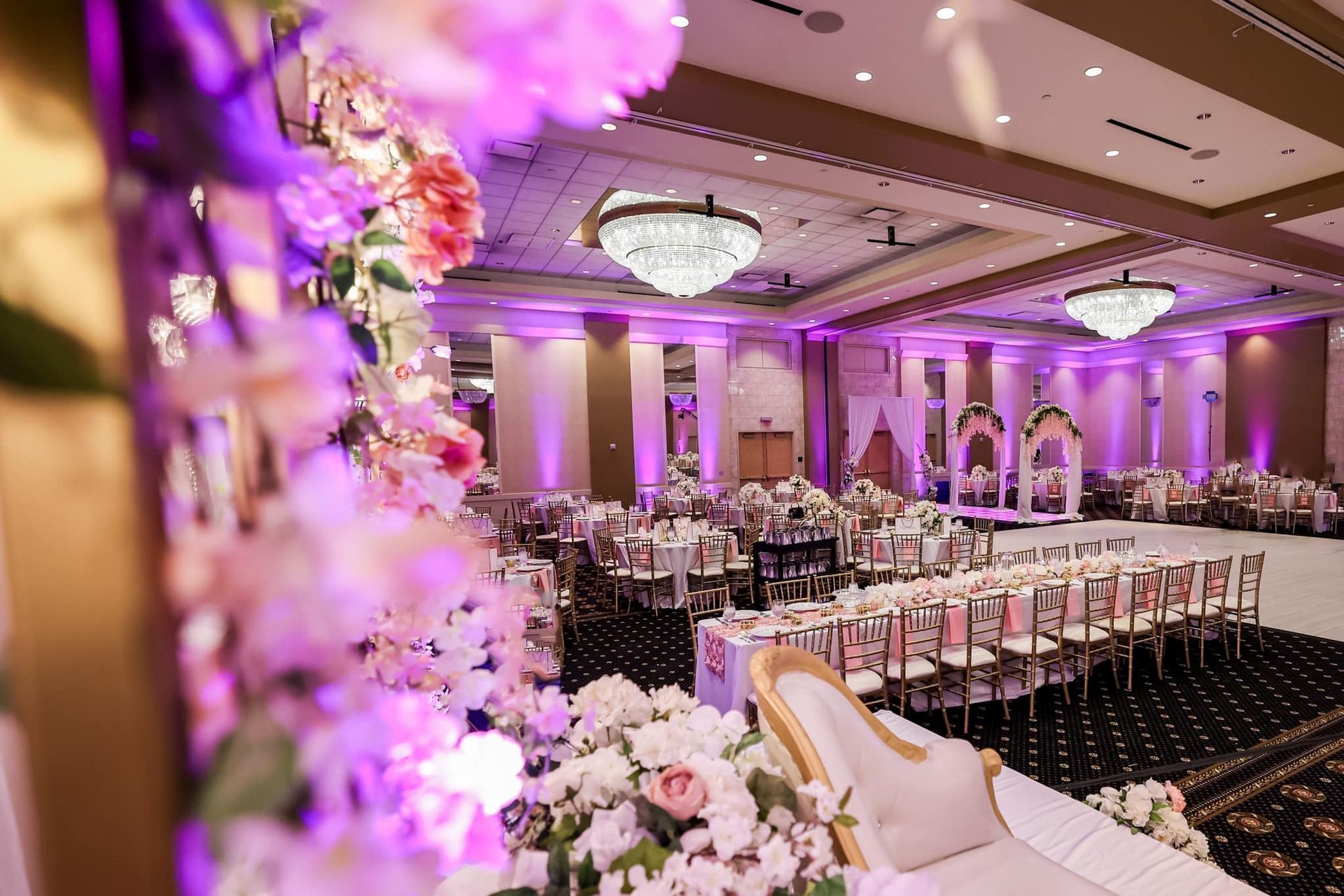 Elegant purple-lit banquet hall with floral decor, round tables, and a white aisle leading to the stage