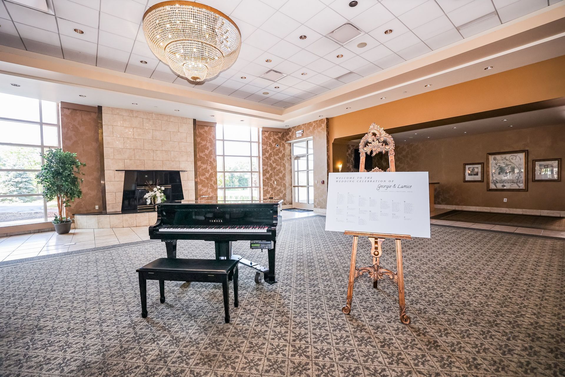 Grand piano in a carpeted ballroom with a sign on an easel and chandelier overhead