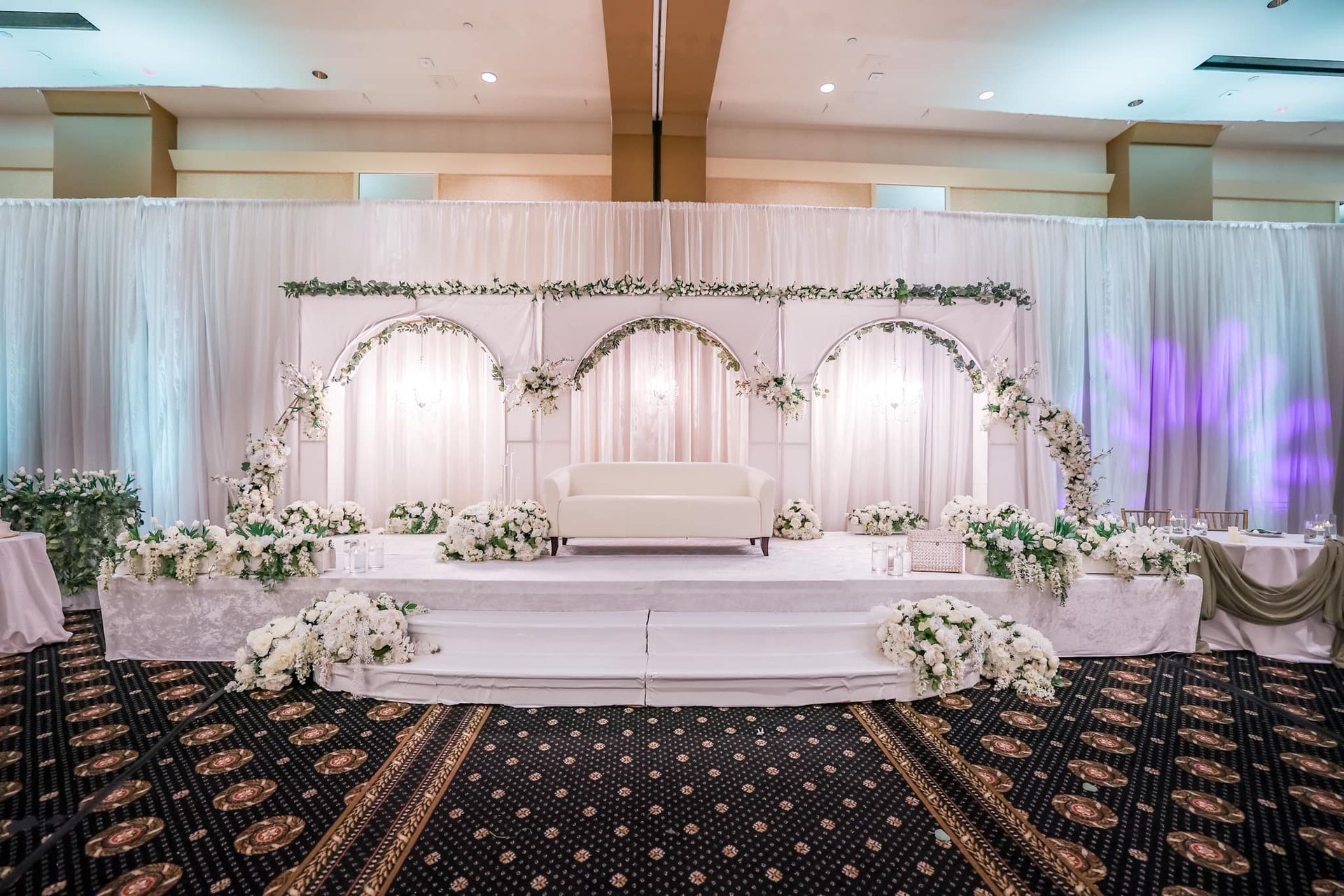 Elegant wedding stage with white drapes, floral arches, and a central loveseat on a patterned carpeted floor