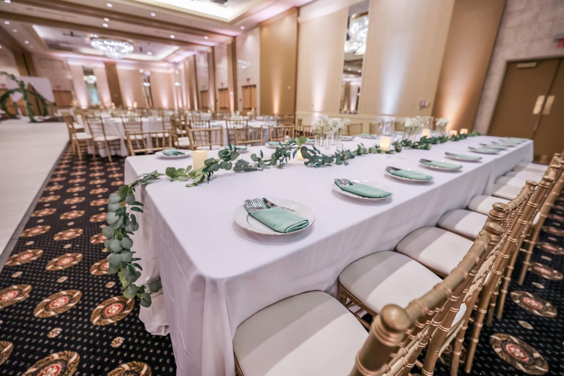 Elegant banquet hall with a long white-clothed table, gold chairs, and green floral garland decorations.