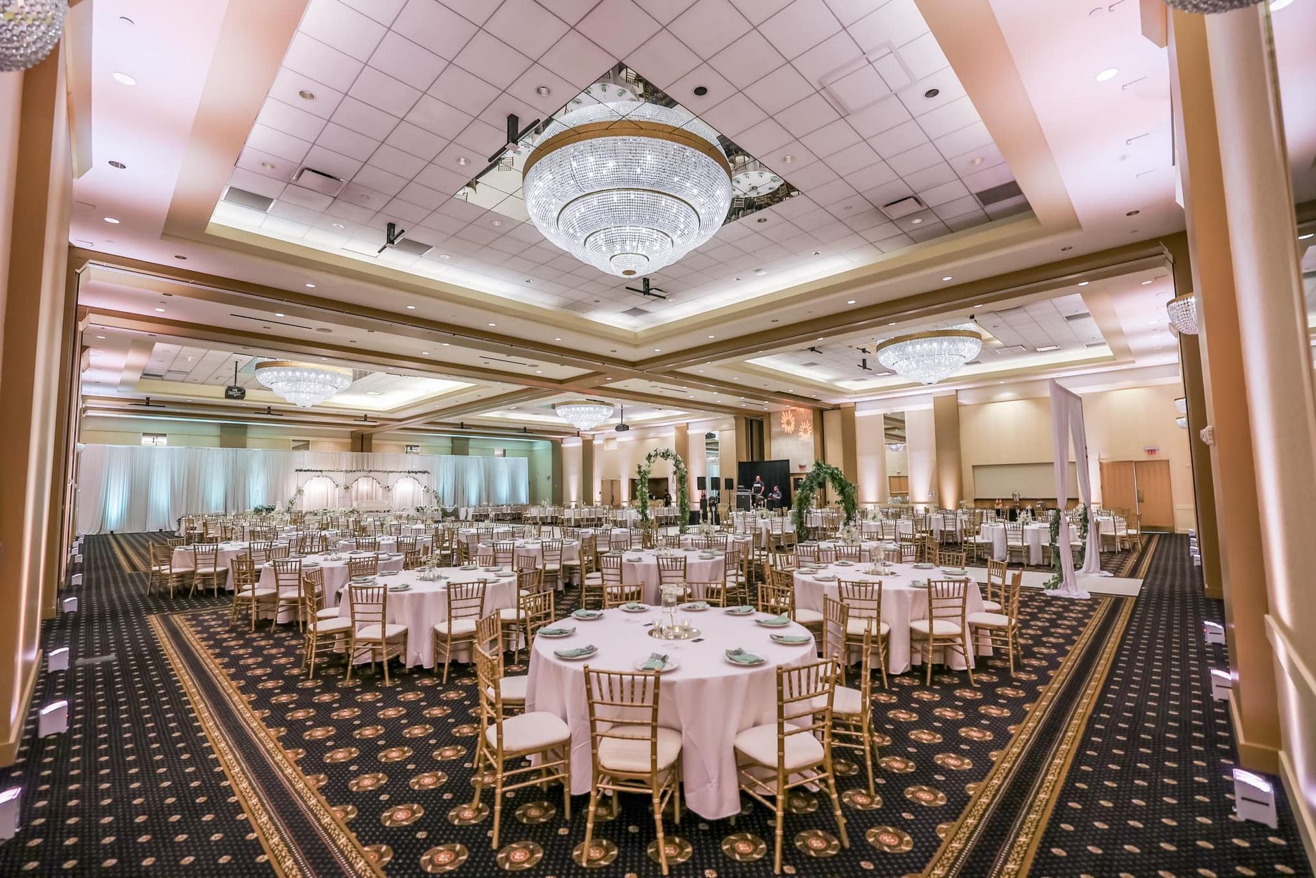 Elegant banquet hall with round tables, white chairs, chandeliers, and patterned carpet prepared for an event