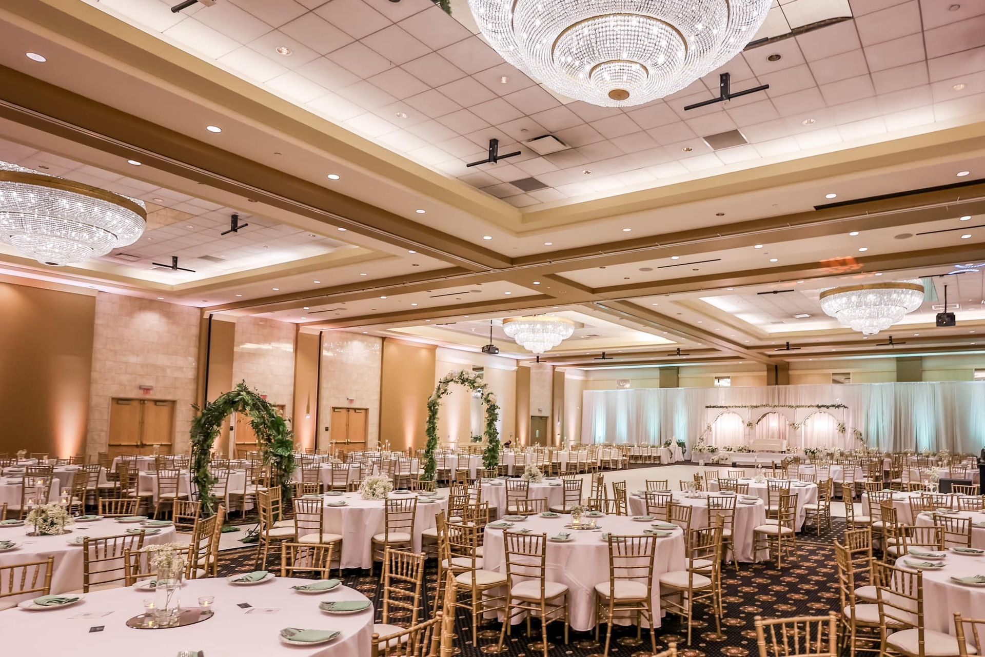 Elegant banquet hall with round tables, gold chairs, chandeliers, and arch decorations set for an event