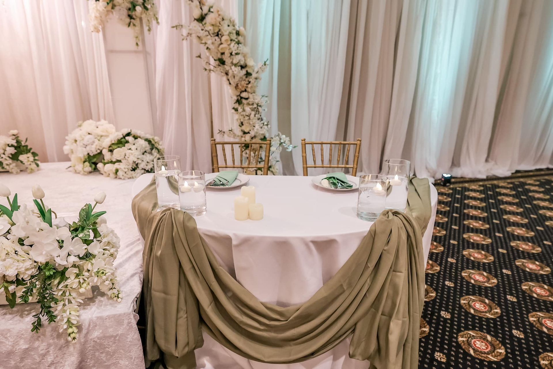 Decorated wedding table with white and olive drapes, floral arrangements, and candles in an indoor reception setting