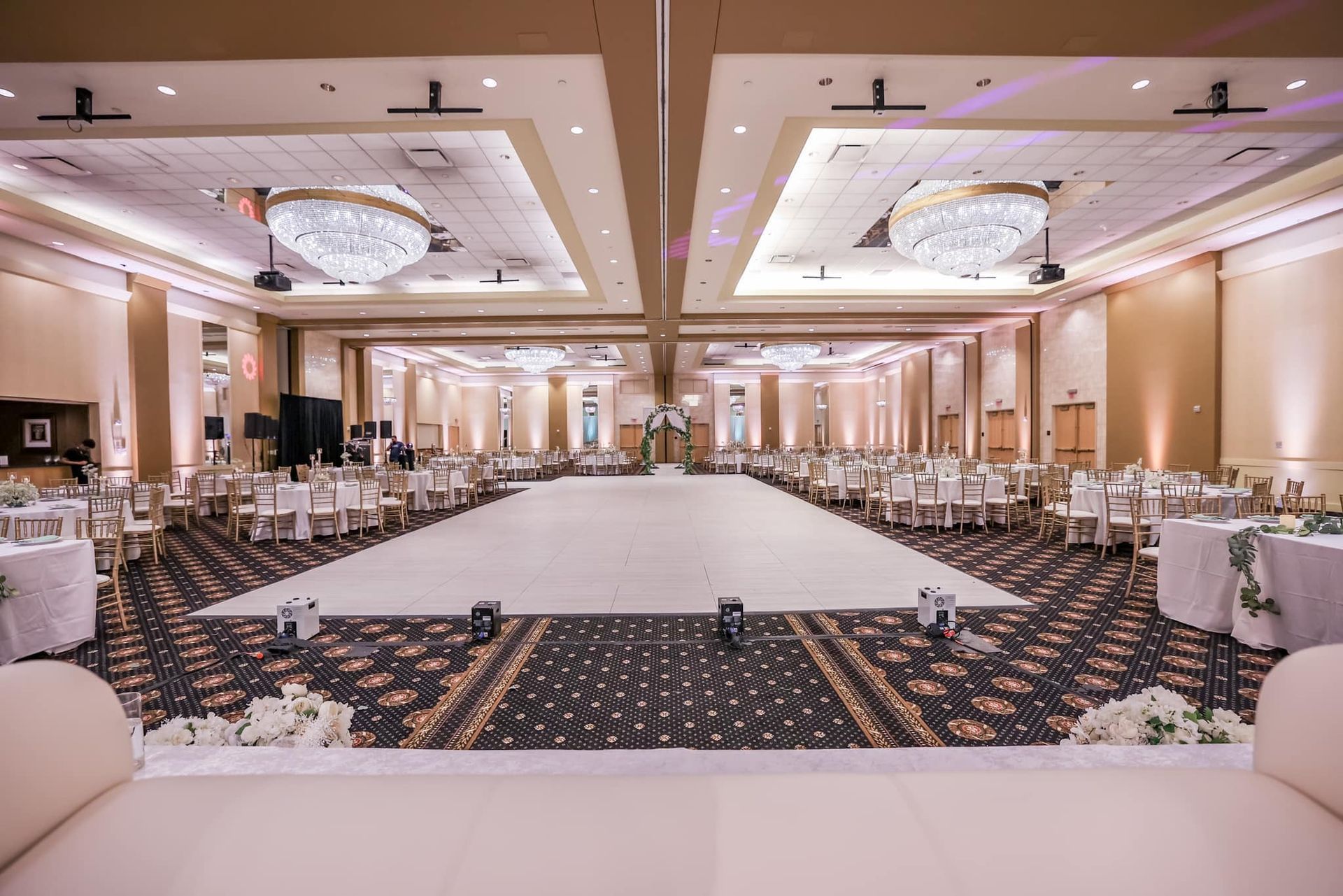 Elegant banquet hall with round tables, white chairs, and a large empty dance floor under chandeliers