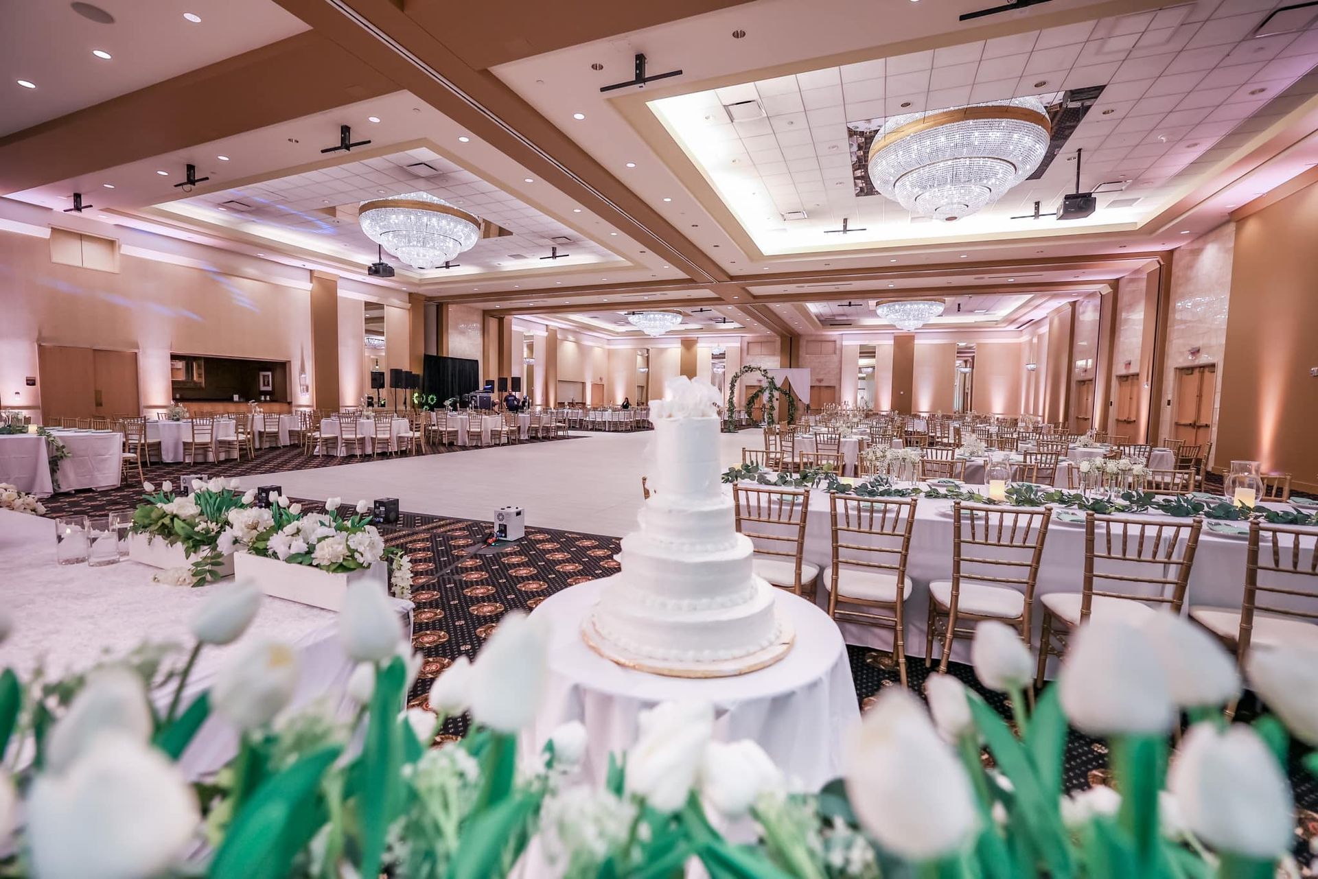 Elegant wedding banquet hall with white tiered cake, floral decor, and rows of chairs under chandeliers