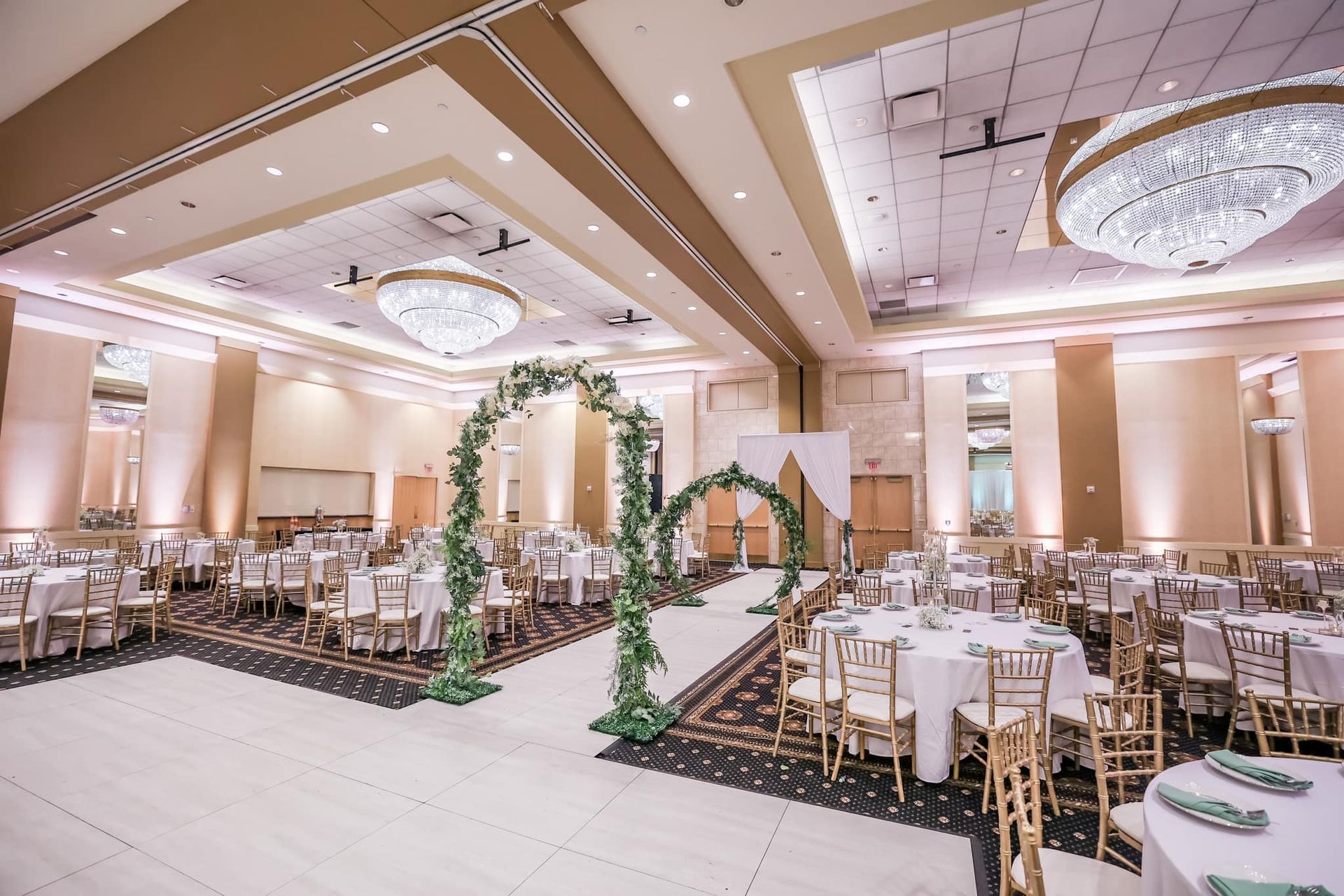 Elegant banquet hall with round tables, gold chairs, chandeliers, and floral arch decor