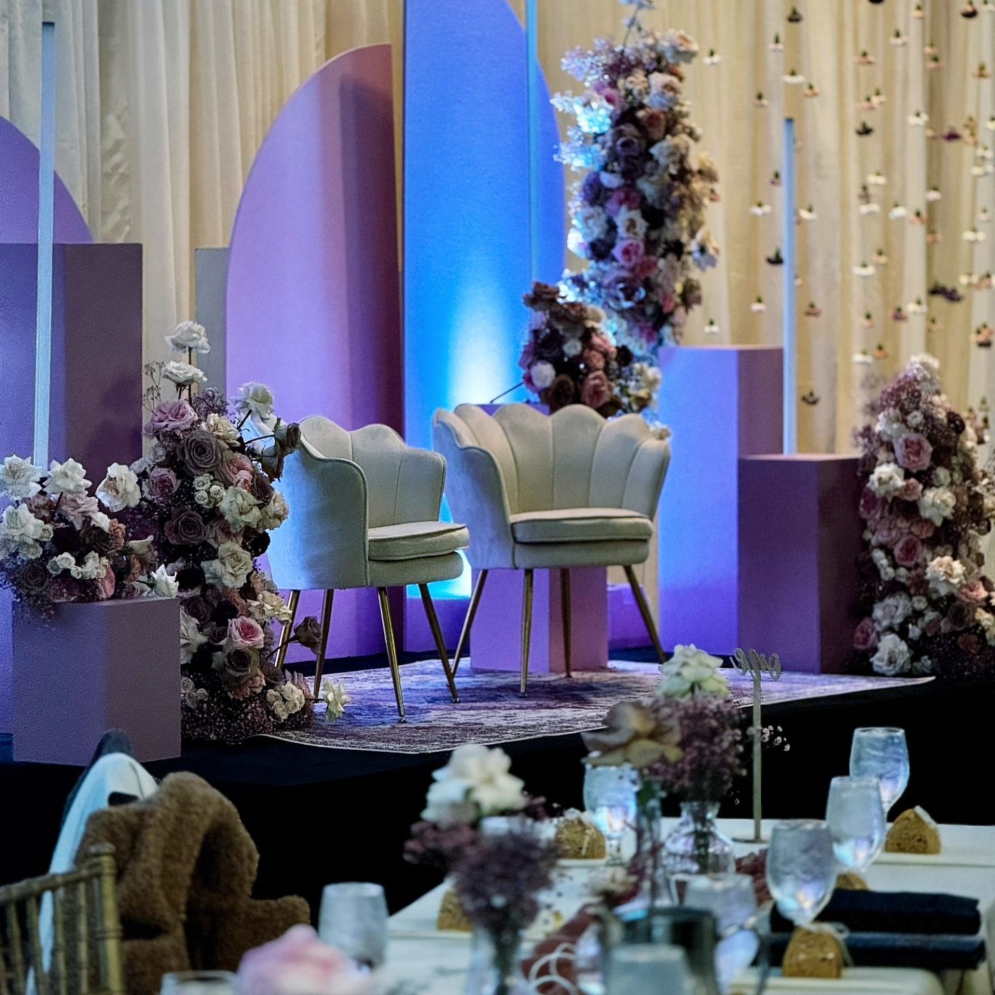 Elegant floral wedding stage with white chairs, purple lighting, and decorated tables in front