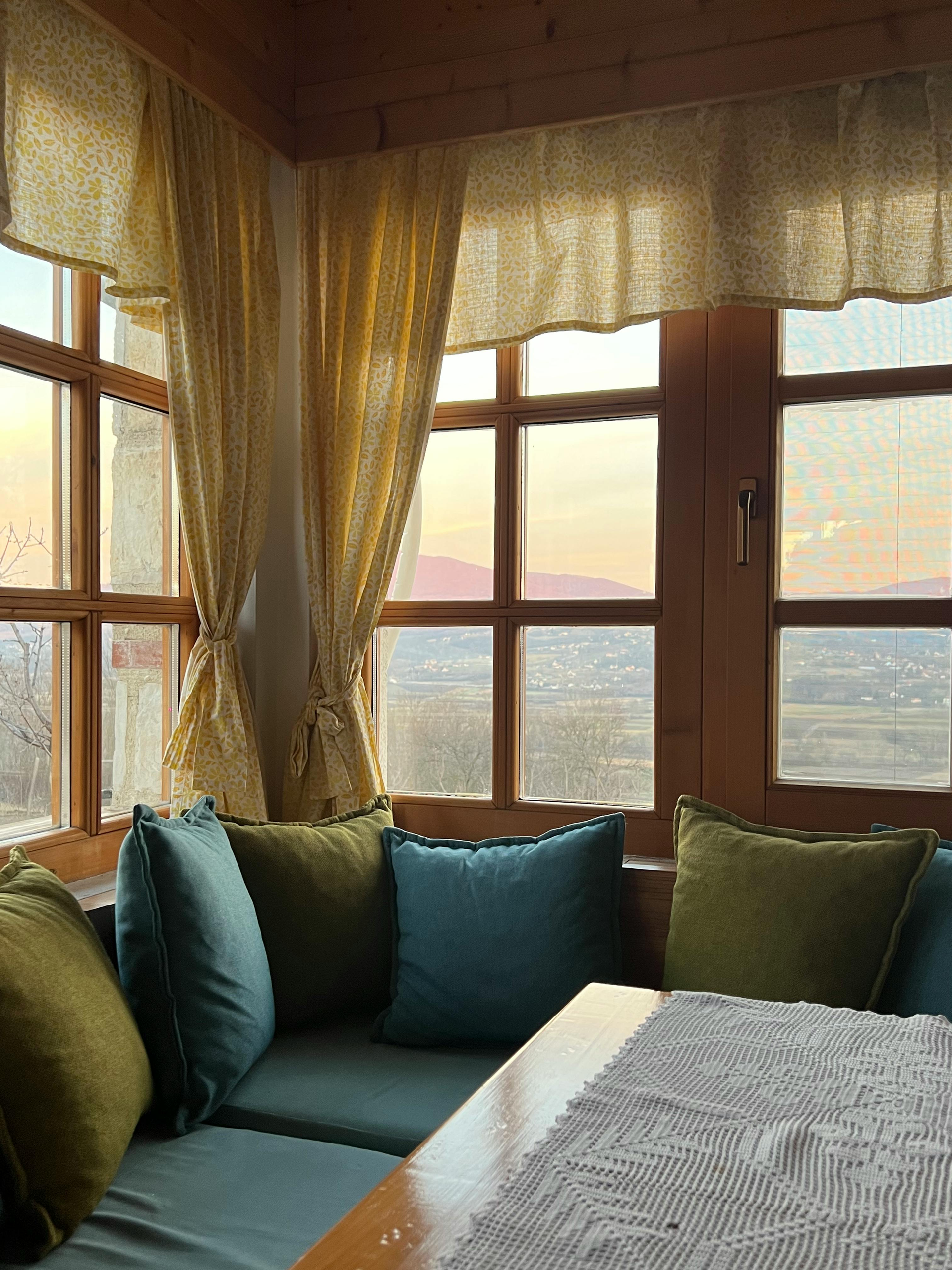 Cozy window seat with blue cushions, gold curtains, and a view of the sea at sunset.