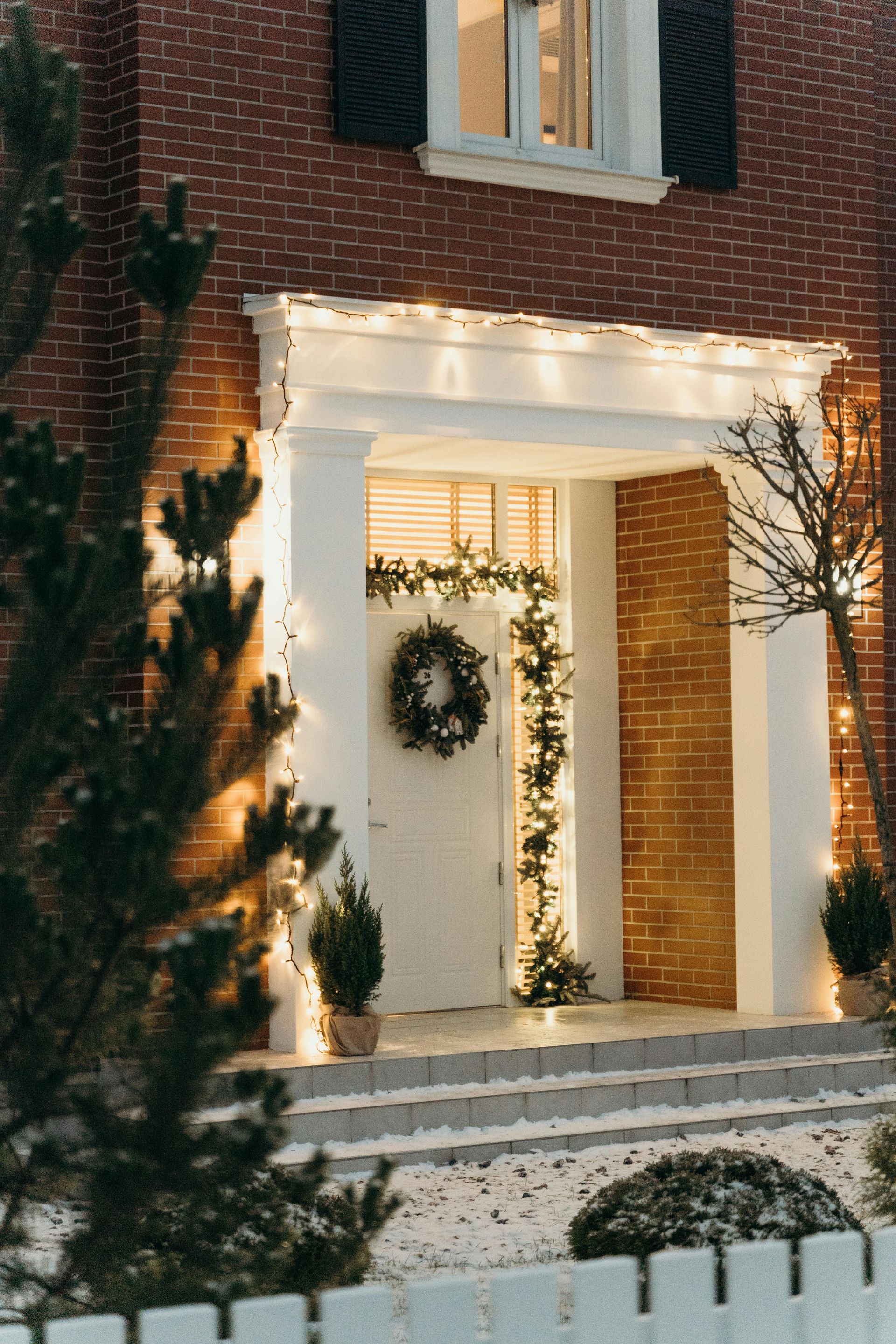 Brick house entry decorated with a holiday wreath, lights, and greenery at dusk