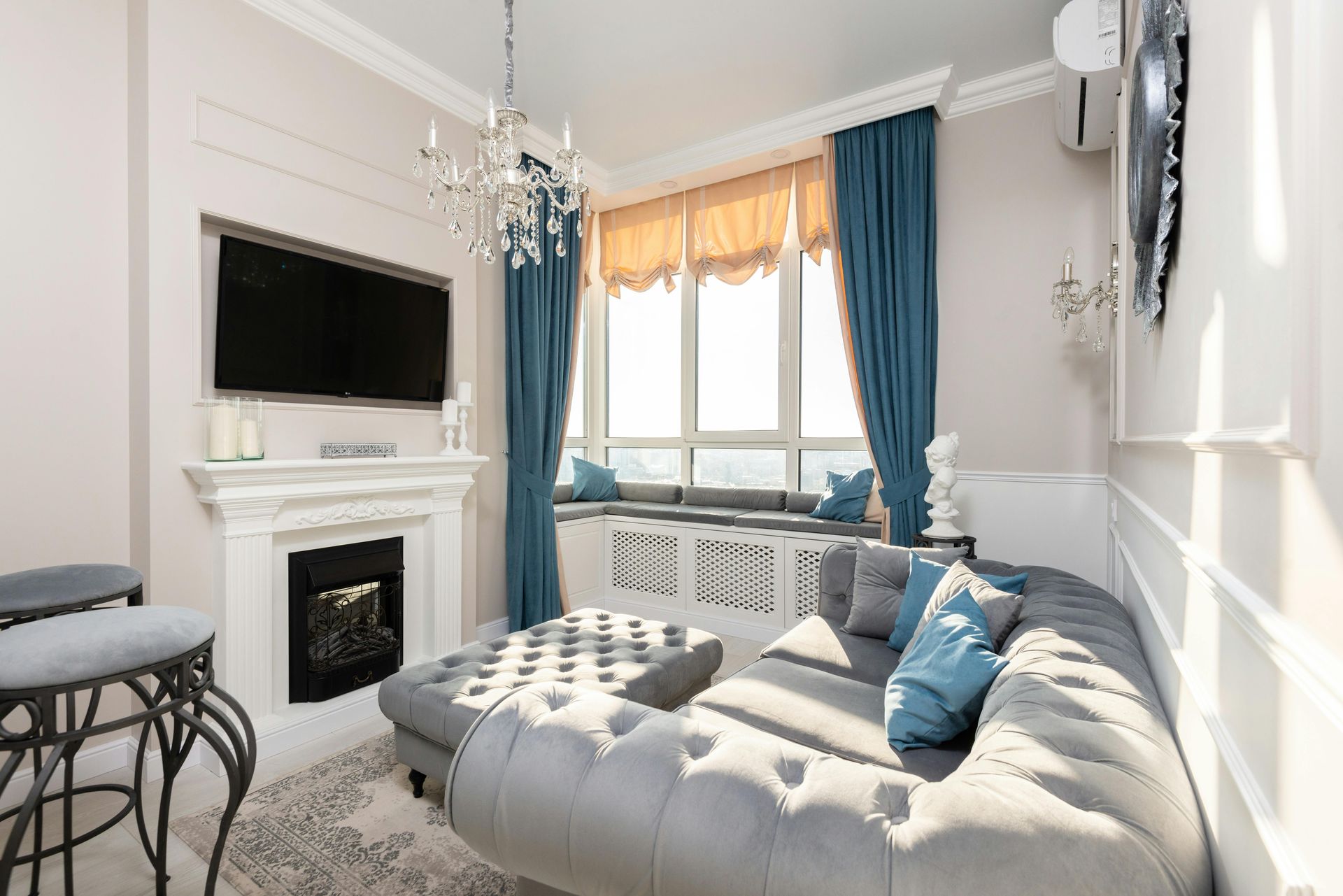 Bright living room with blue curtains, tufted gray chaise, fireplace, TV, and window seating