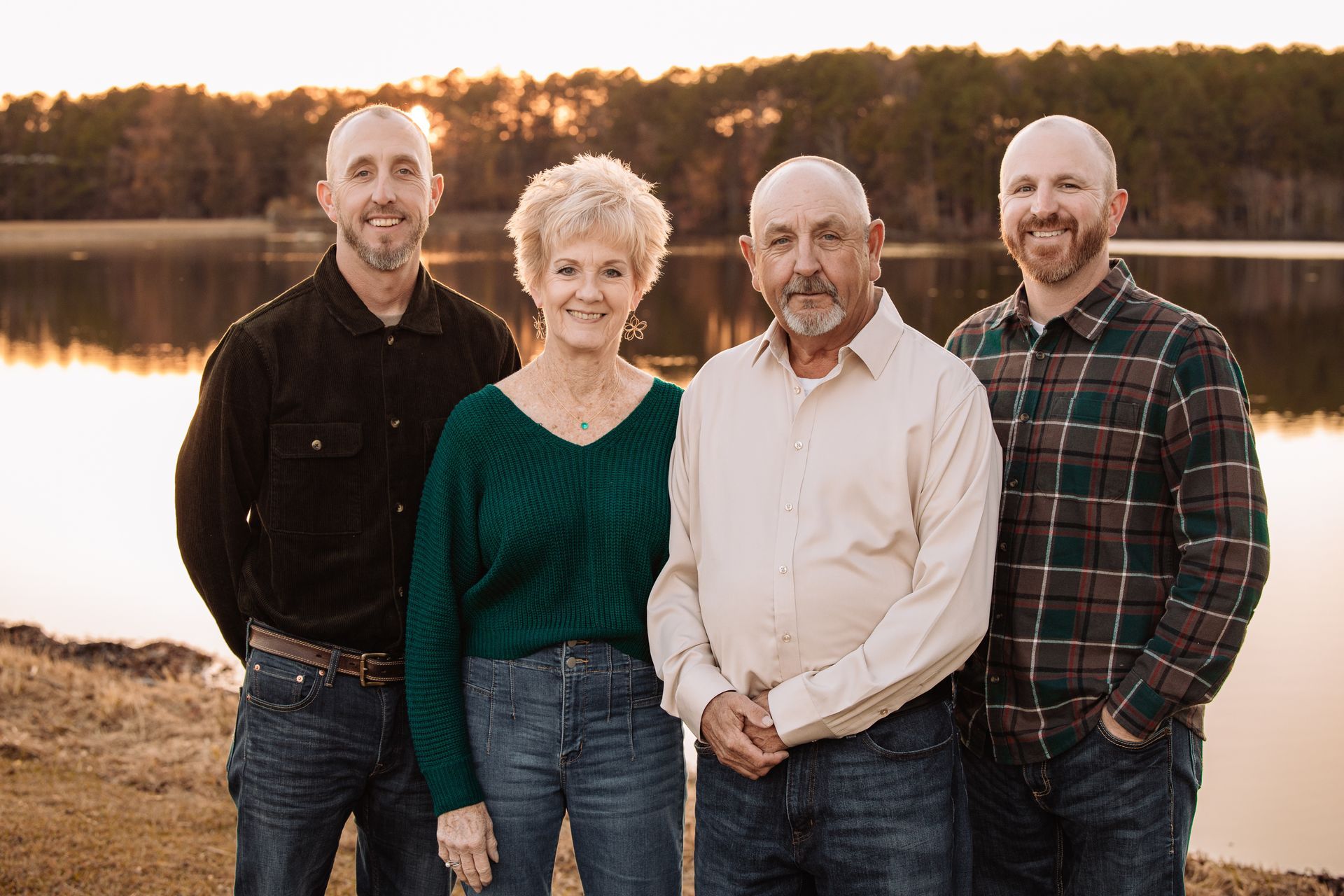 Four people standing together by a lake at sunset, smiling in casual clothes