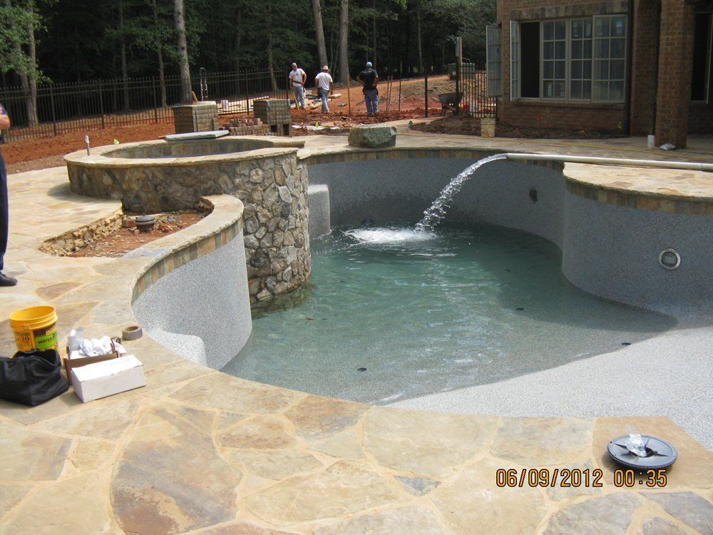 Backyard pool under construction with stone walls, flowing water, and workers in the background