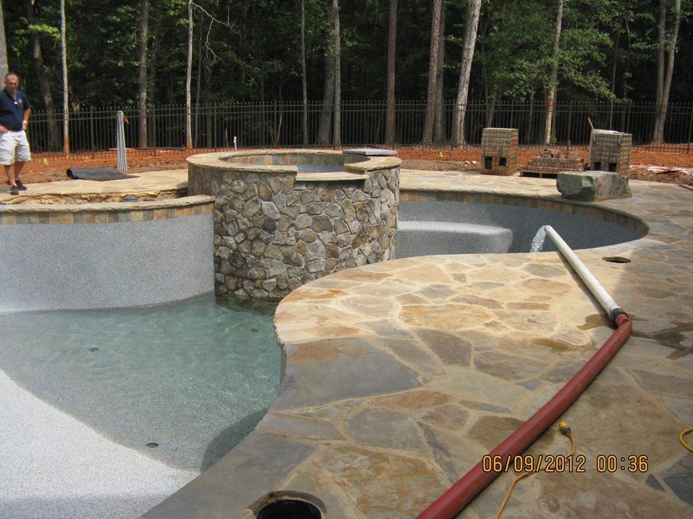 Outdoor stone splash pad with circular water basins, pipes, and a wooded background