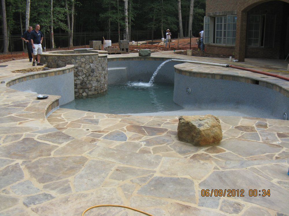 Stone patio around a small pool with a waterfall, in a wooded backyard.